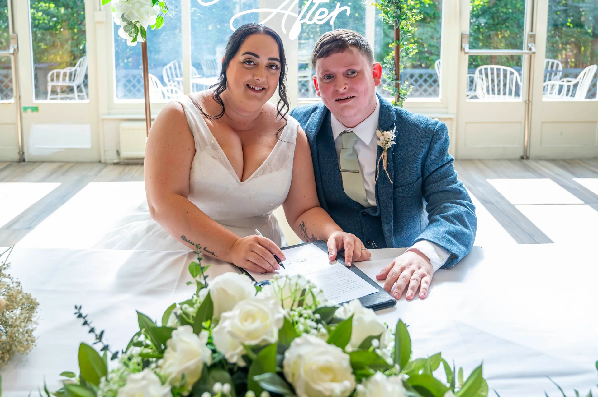 The bride and groom are smiling at the camera while signing the marriage register at a table adorned with white roses and greenery.