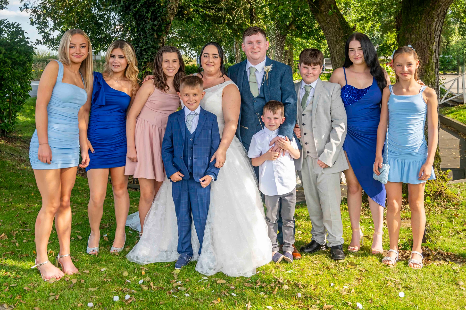 A large wedding party poses outdoors on a grassy lawn with trees in the background, featuring the bride in a white gown and the groom in a blue suit surrounded by family and bridesmaids.