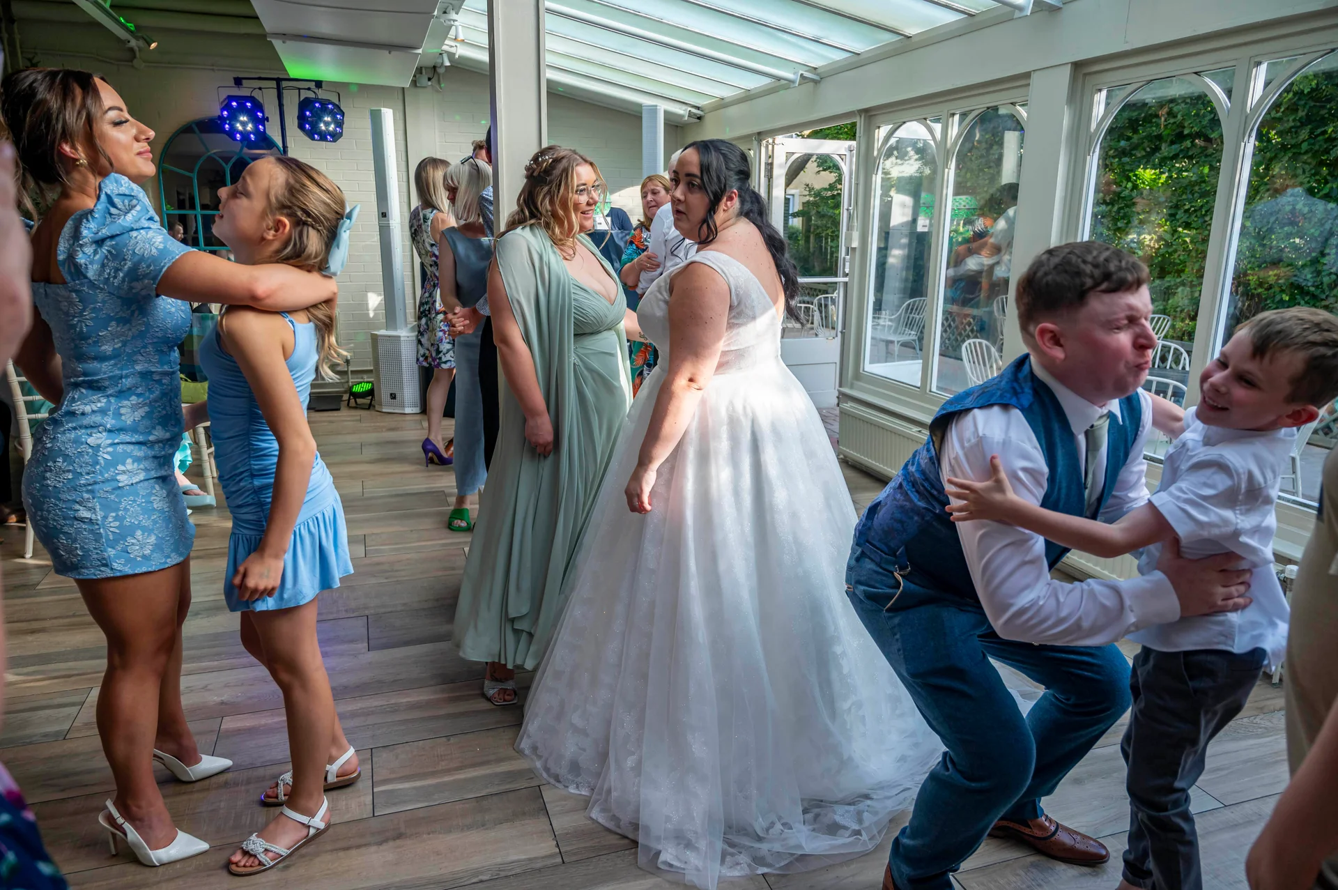 A candid wedding reception scene inside a glass-roofed conservatory featuring a bride in a sparkling a-line gown and guests dancing and laughing.