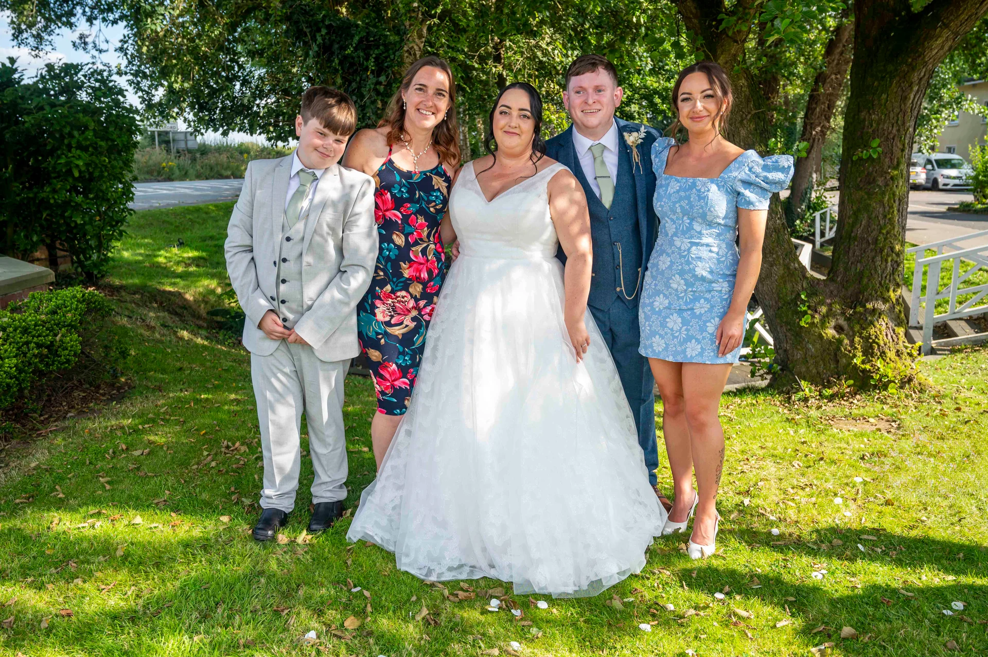 A happy wedding party poses outdoors on a sunlit lawn next to trees, featuring the bride in a white A-line gown, the groom in a blue suit, and guests in coordinated attire.