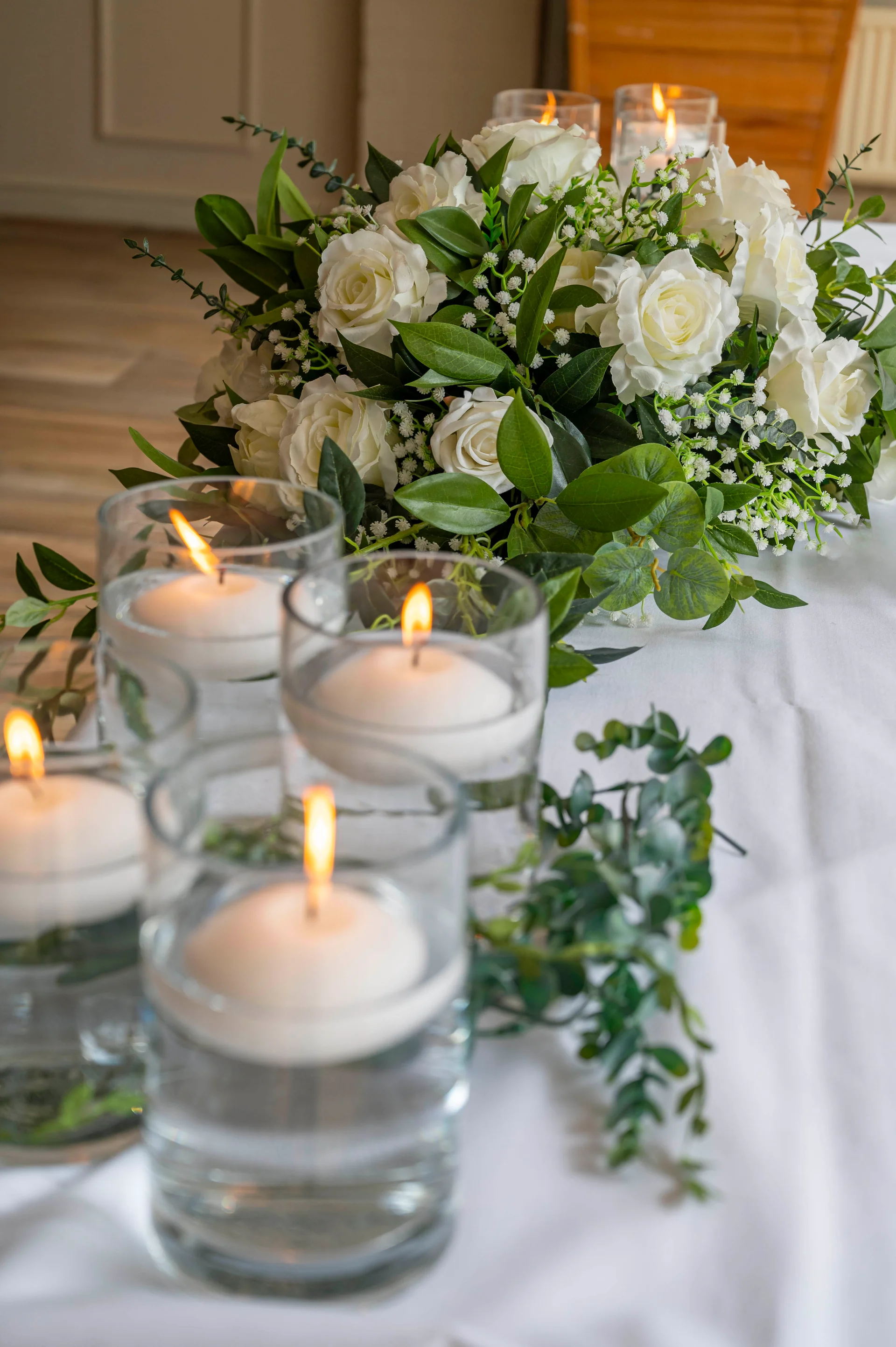 A close-up of a wedding reception table featuring floating candles in glass cylinders alongside a lush arrangement of white roses and green foliage.