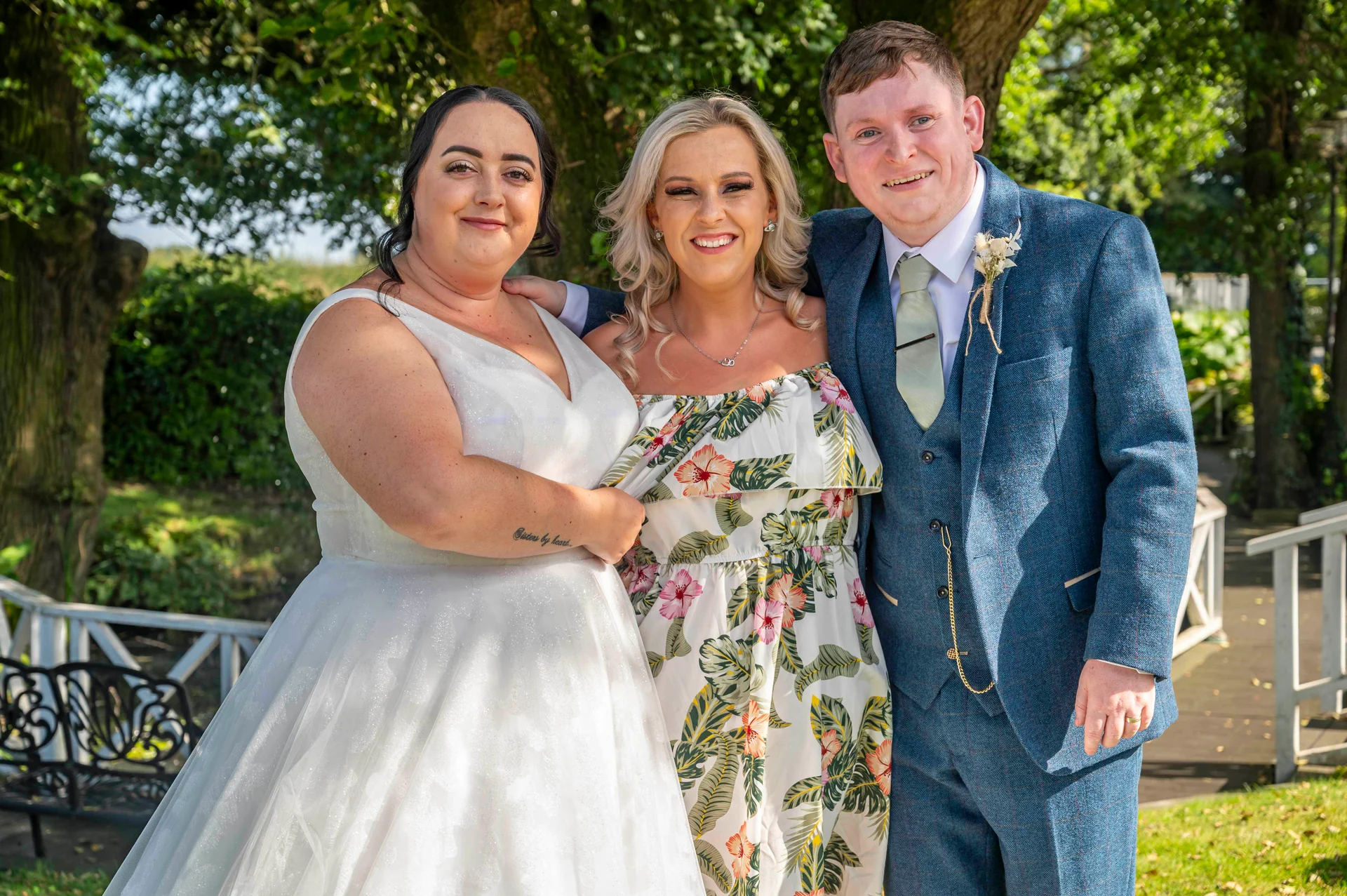 A smiling bride in a white wedding gown stands alongside a groom in a blue tweed suit and a guest in a floral dress outdoors in a sunny garden.