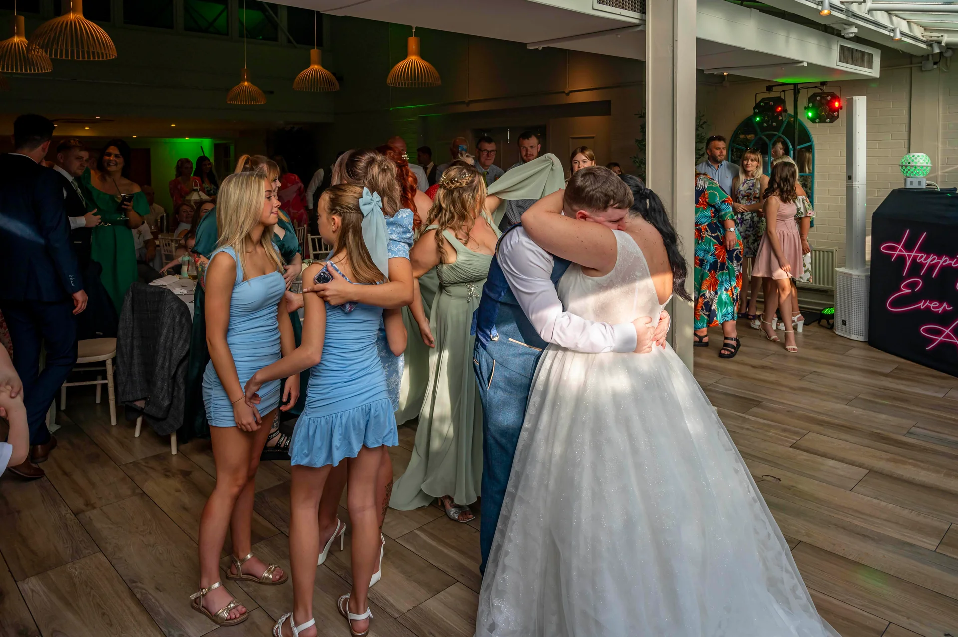 The newlywed couple share a tight embrace on the dance floor during their evening reception, surrounded by guests watching in a brightly lit venue.