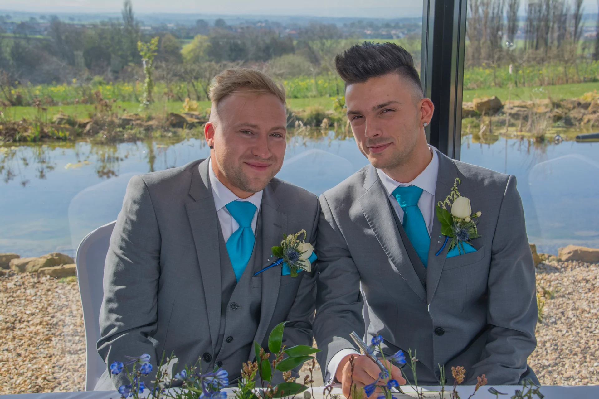 Two grooms in matching grey three-piece suits and vibrant turquoise ties smile as they sit together by a window with a scenic lakeside view during their wedding ceremony.