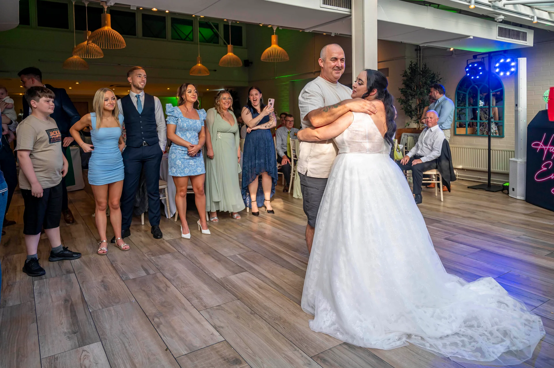 A bride in a white sparkling a-line gown shares a dance with a family member while wedding guests watch from the sidelines.