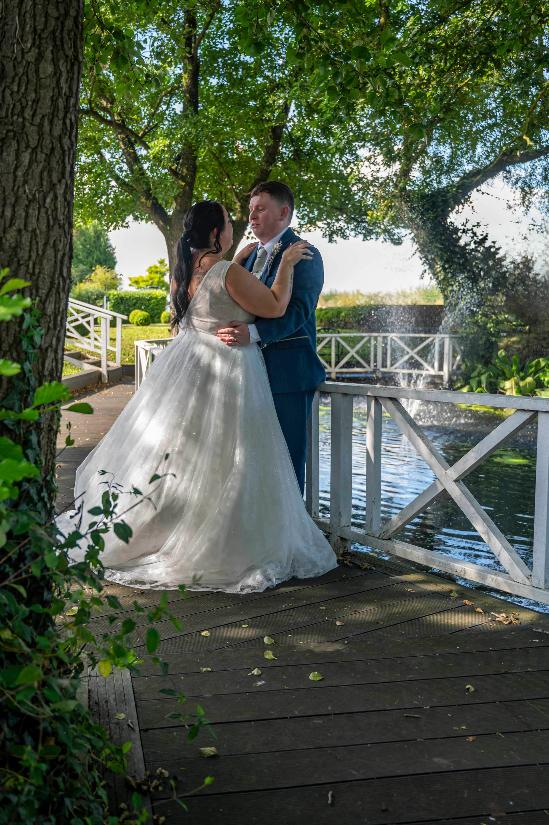 A newlywed couple stands embracing on a wooden footbridge over a pond, surrounded by lush trees and a sparkling fountain in the background.