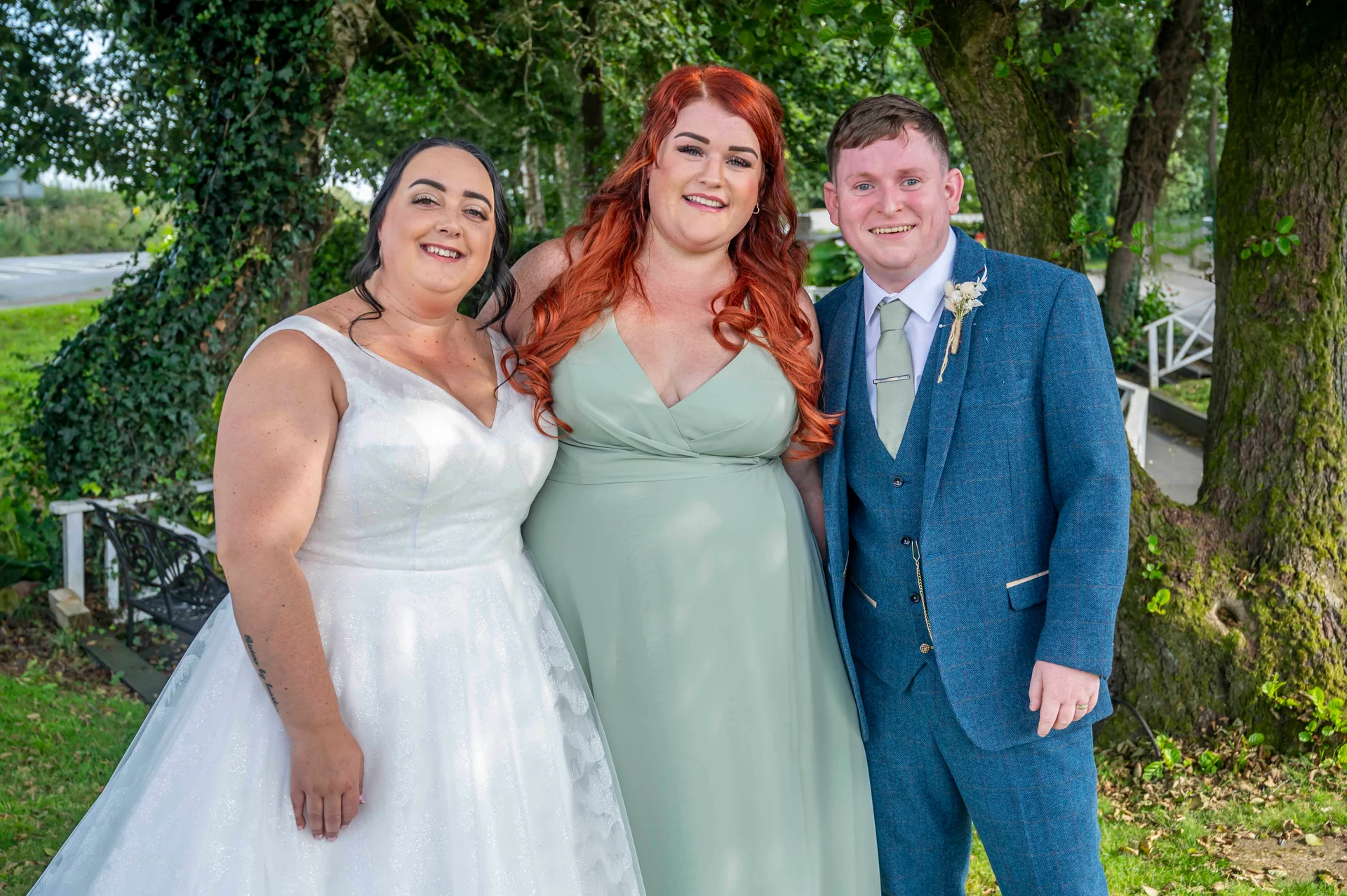 A smiling bride in a white gown and a groom in a blue tweed suit pose outdoors next to a bridesmaid wearing a sage green dress under leafy trees.