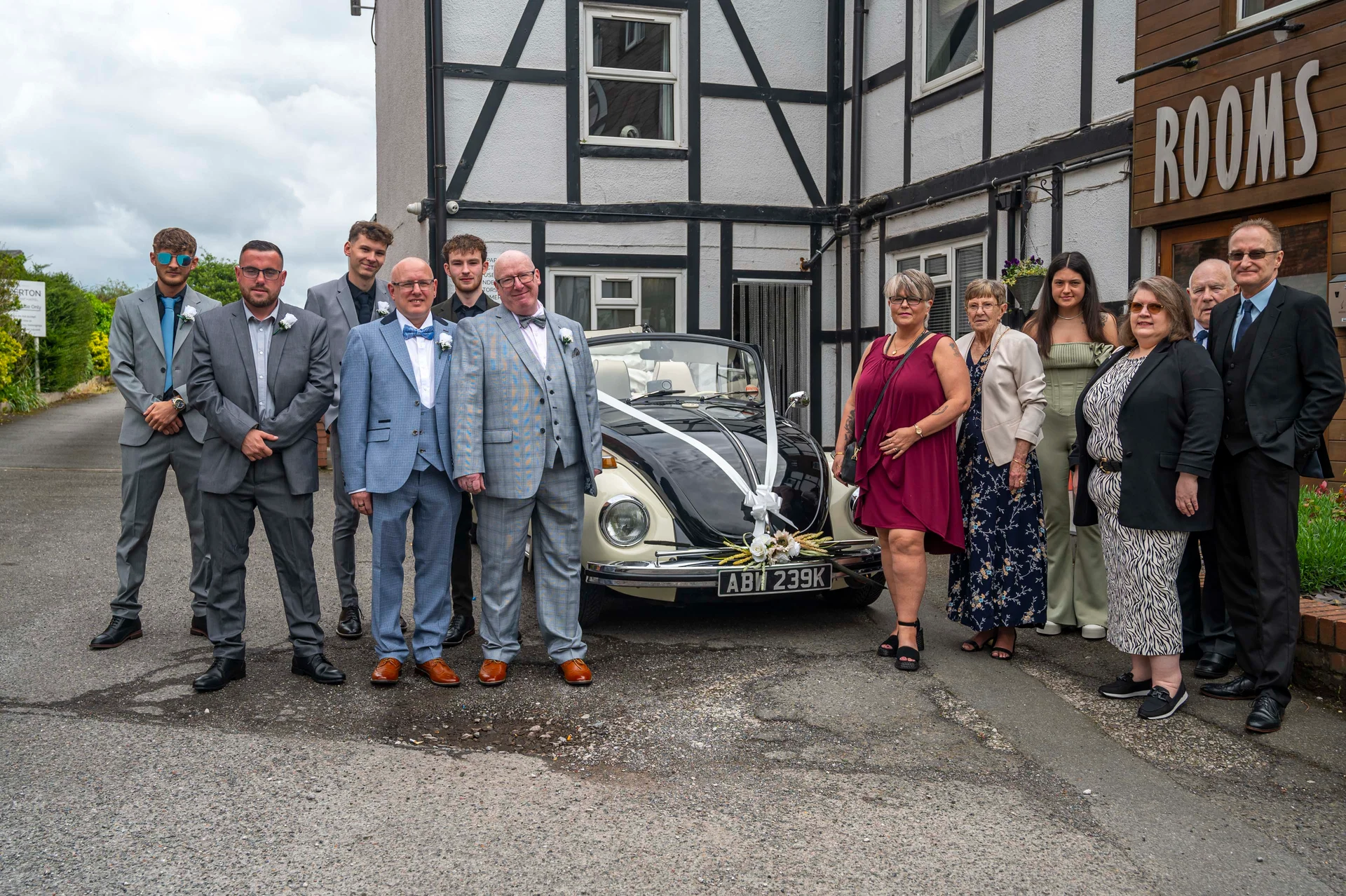 A group of family members and groomsmen pose together in front of a vintage convertible Volkswagen Beetle decorated with white ribbons outside a traditional building.