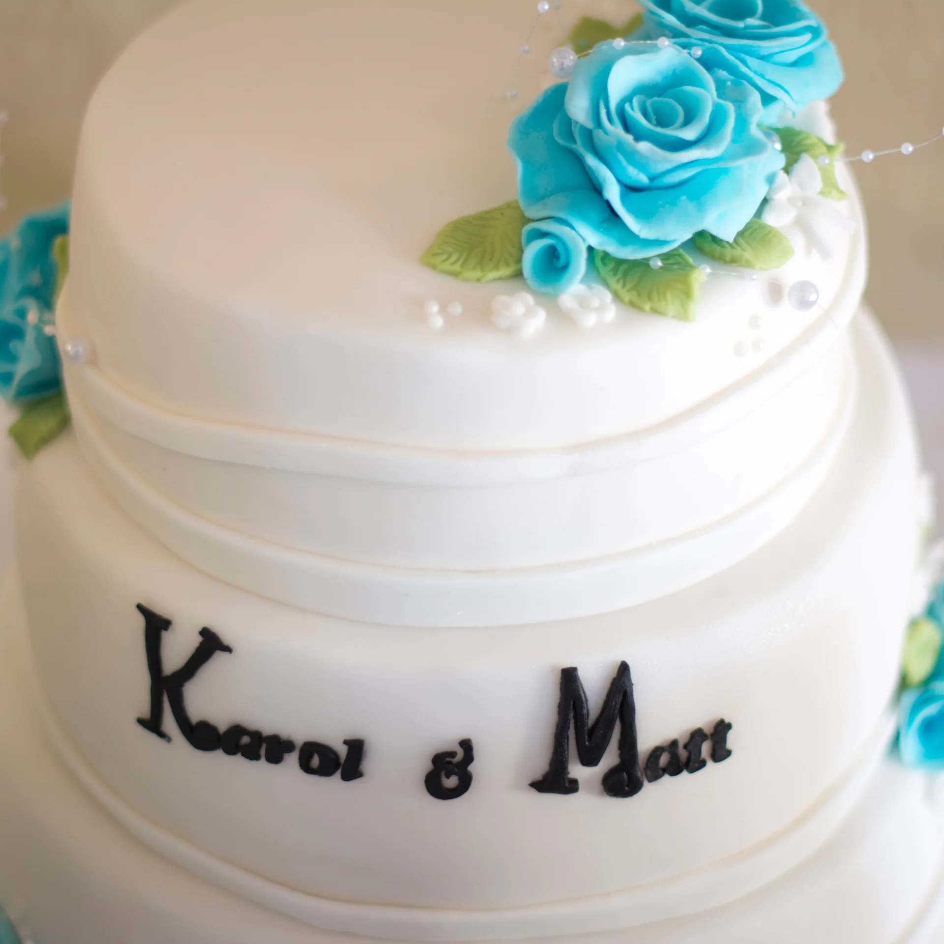 A close-up shot of a white multi-tiered wedding cake adorned with bright blue sugar roses and personalised black lettering on the lower tier.