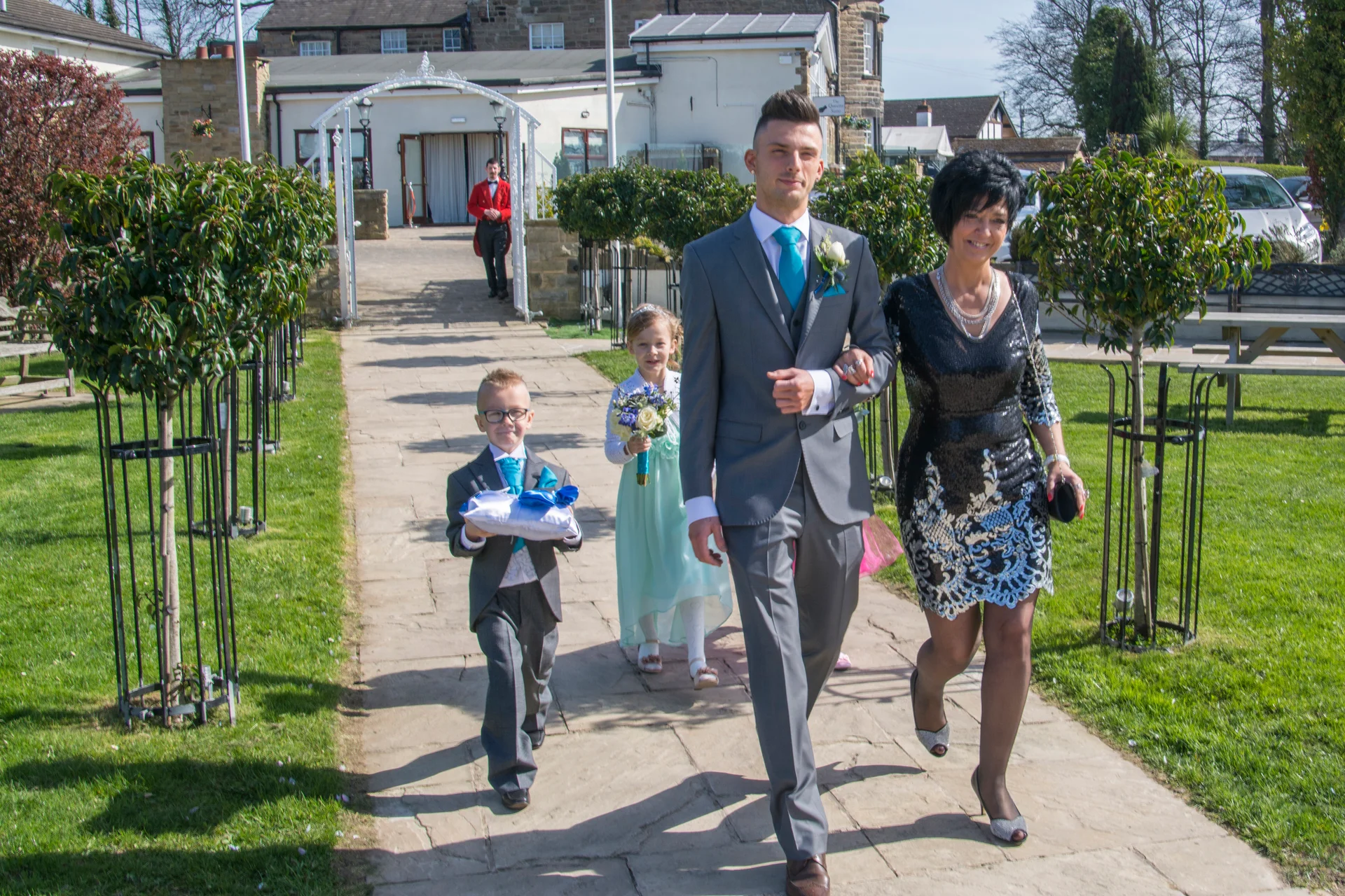 A groom in a grey suit and bright blue tie walks down a paved outdoor aisle alongside family members, including a page boy carrying a ring pillow and a flower girl holding a bouquet.