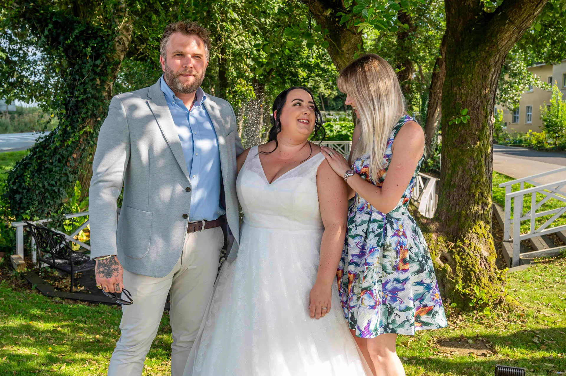 A groom in a grey jacket and open-collared shirt stands beside a bride in a white A-line gown while a guest in a floral dress looks on in a sunlit garden.