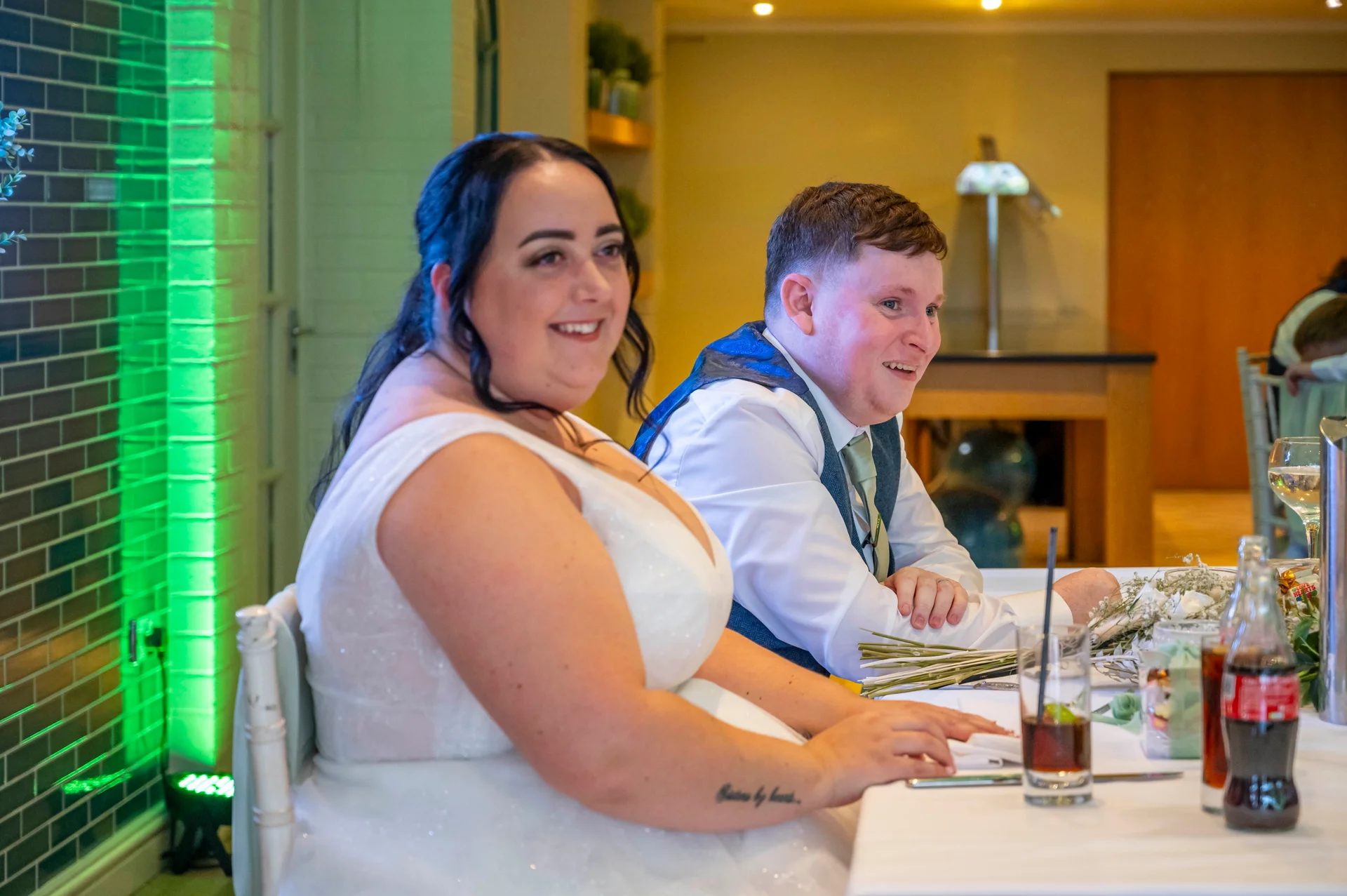 A smiling bride in a white sleeveless gown and groom in a waistcoat sit together at the top table during wedding speeches.