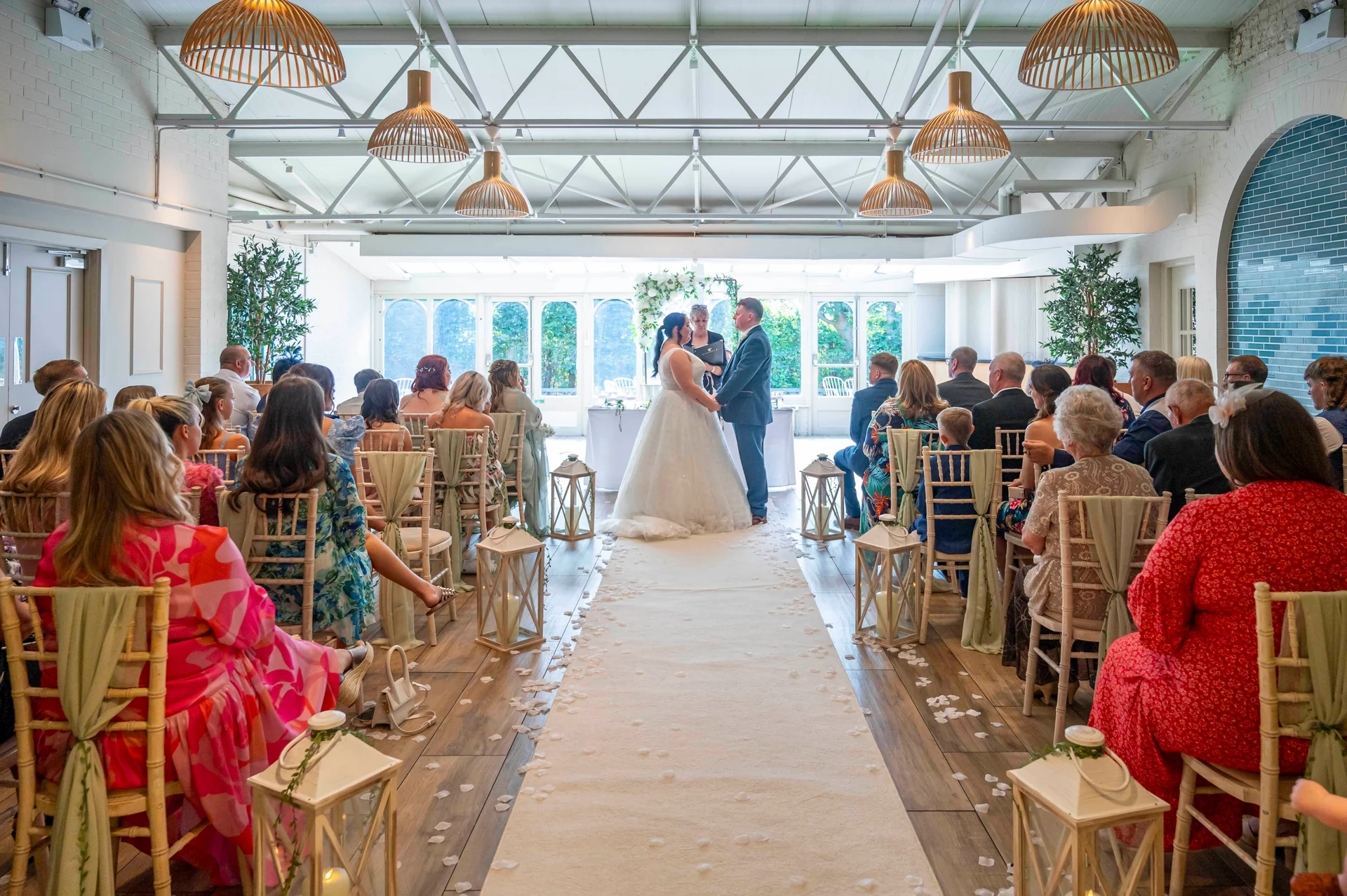 A bride and groom exchange vows at the top of a white aisle runner during a light-filled warehouse wedding ceremony.