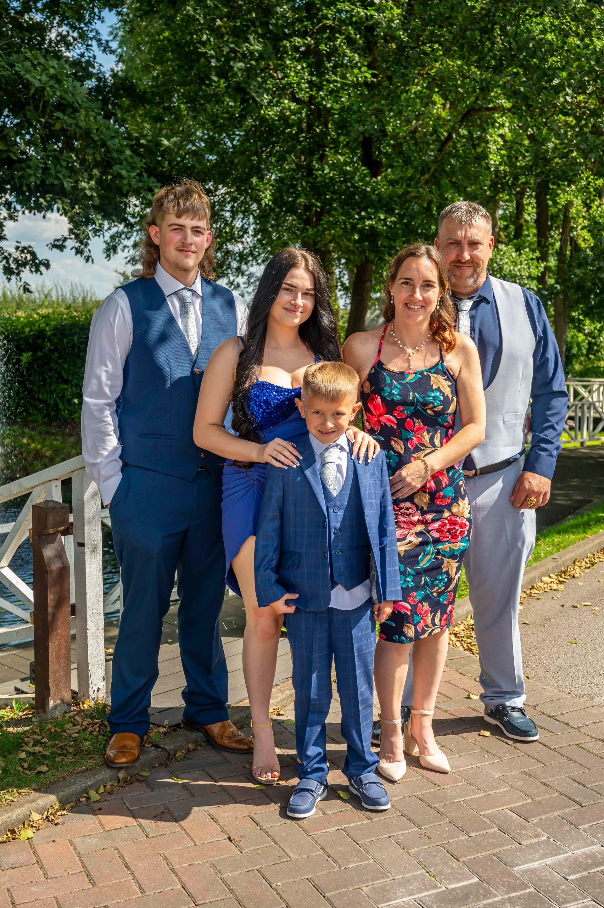 A family group poses outdoors on a paved pathway near a wooden bridge during a sunny wedding celebration.