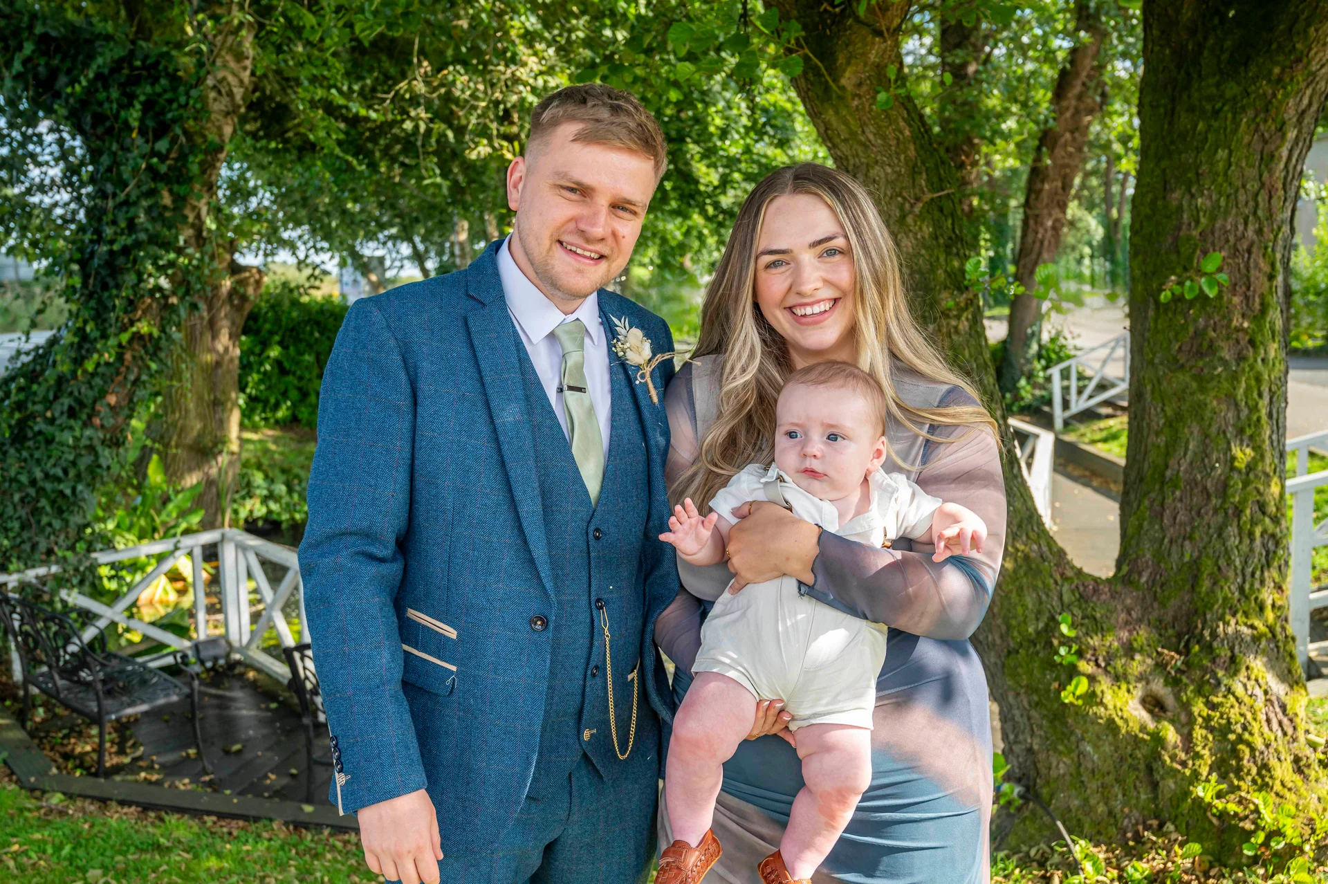 A smiling family portrait featuring a groom in a blue tweed suit, a partner, and a baby standing outdoors near leafy trees.