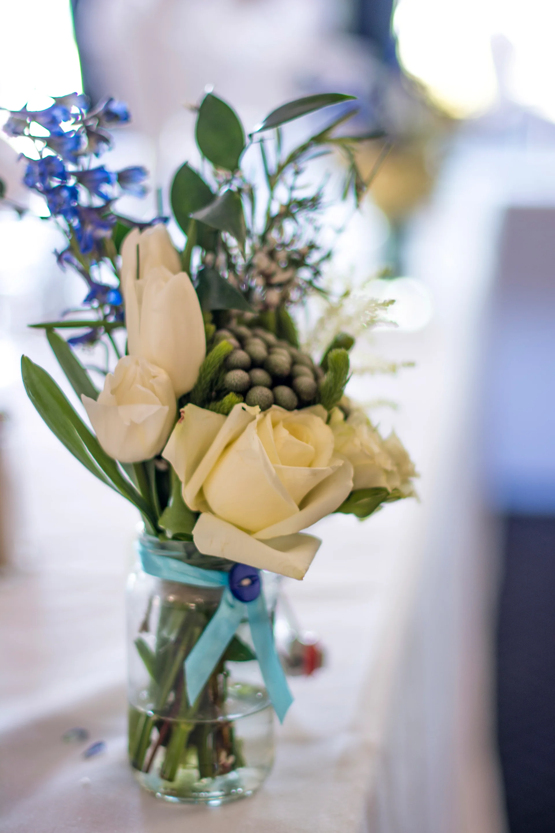 A close-up shot of a delicate table arrangement featuring white roses, tulips, and bluebells in a glass jar tied with a blue ribbon.