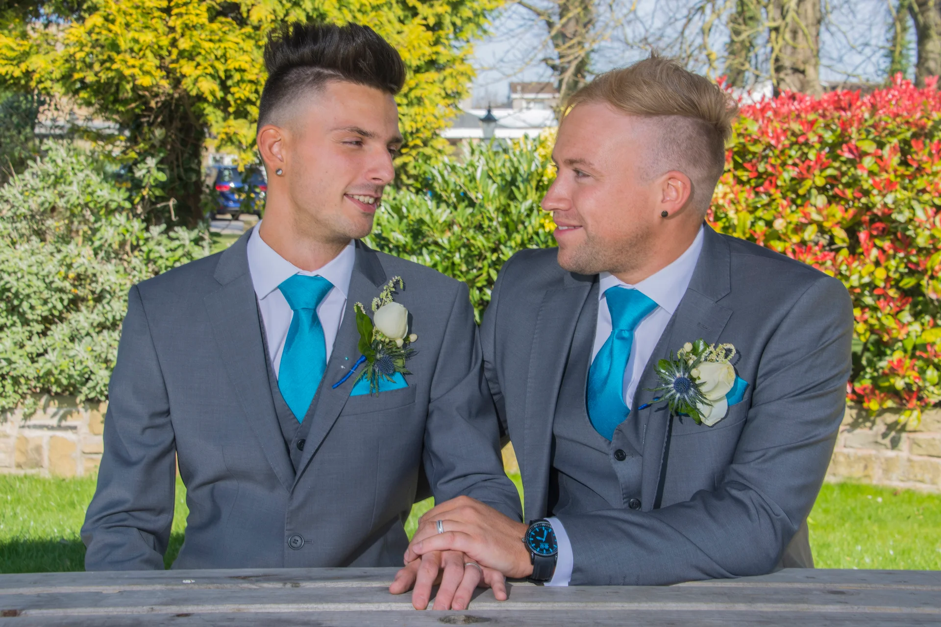 Two grooms in matching grey three-piece suits and turquoise ties hold hands and exchange fond glances while seated outdoors in a sunlit garden.