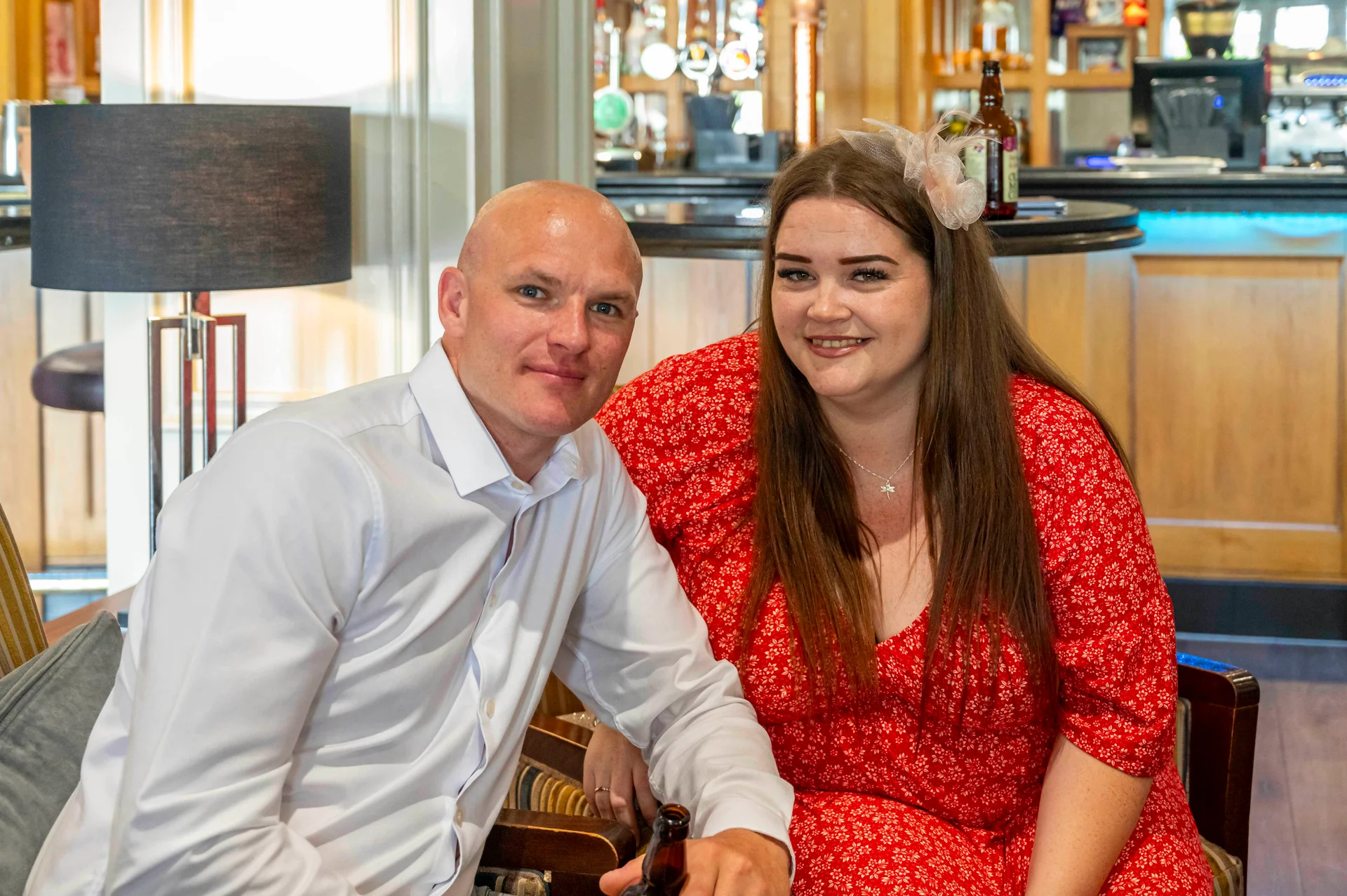 A smiling couple pose together inside a pub during a wedding celebration, with the woman wearing a red floral dress and a fascinator.