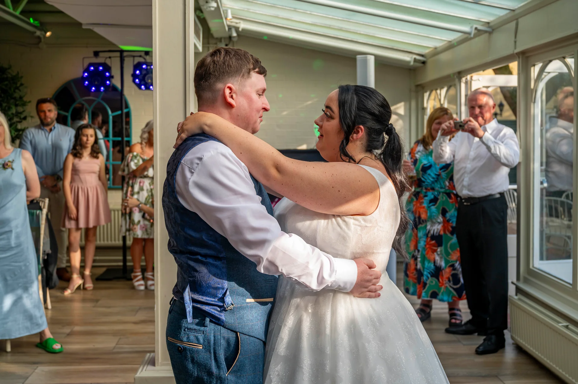 Newlyweds share an intimate first dance together inside a bright conservatory while guests look on and capture the moment.