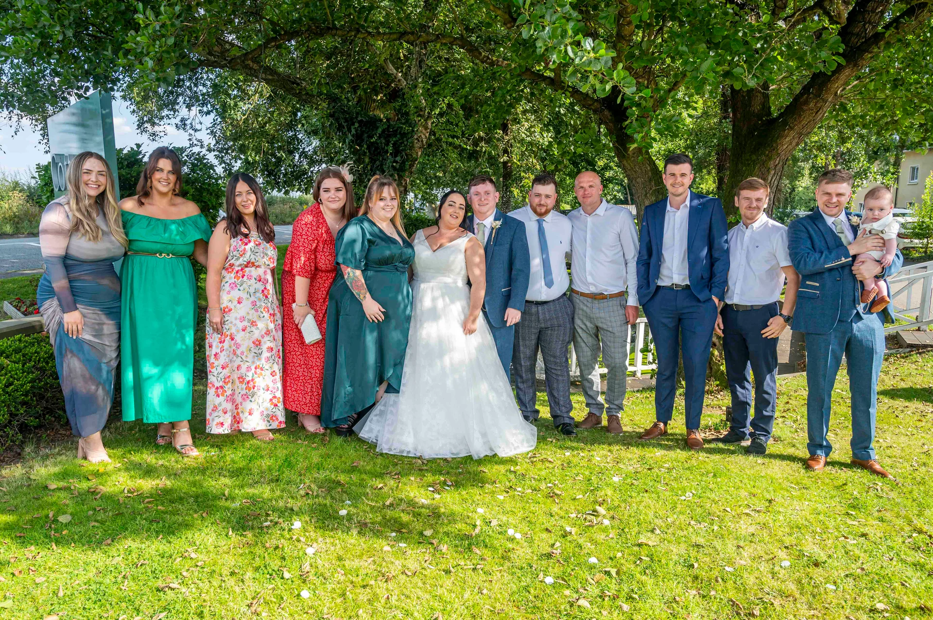 A large wedding party and family group pose together outdoors on a sunny lawn beneath the shade of lush green trees.