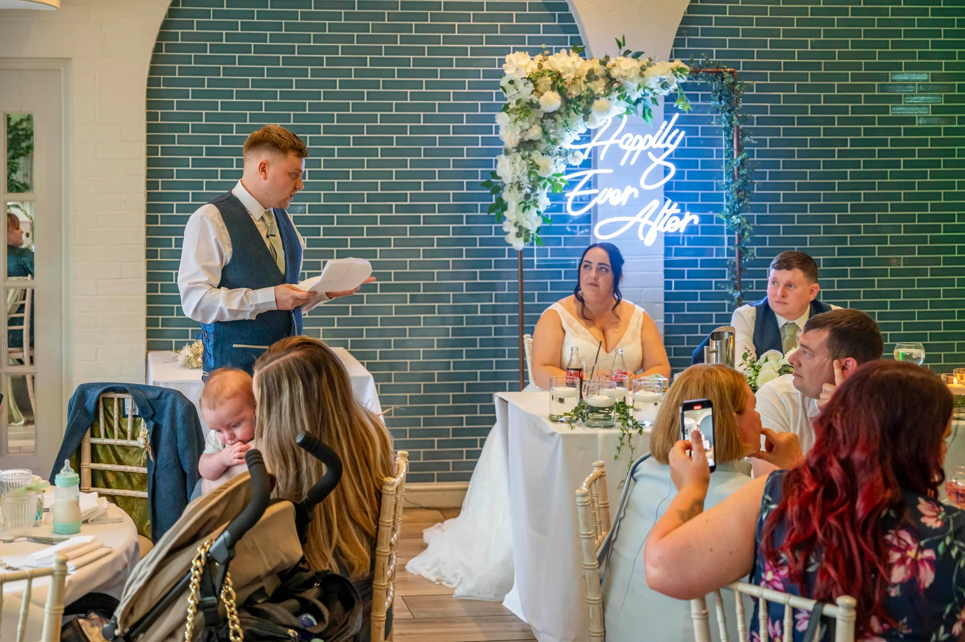 A groom delivers a speech while holding notes during the wedding breakfast, with the bride and guests watching against a striking teal tile and neon sign backdrop.