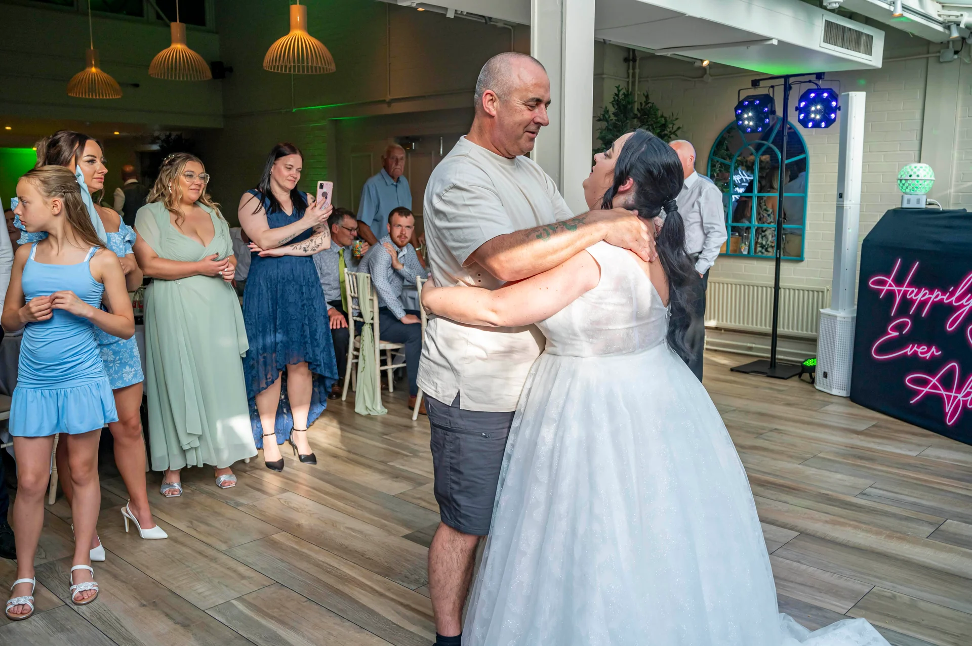 A bride in a white tulle wedding dress shares her first dance with her partner on a wooden dance floor while guests watch and record on phones.