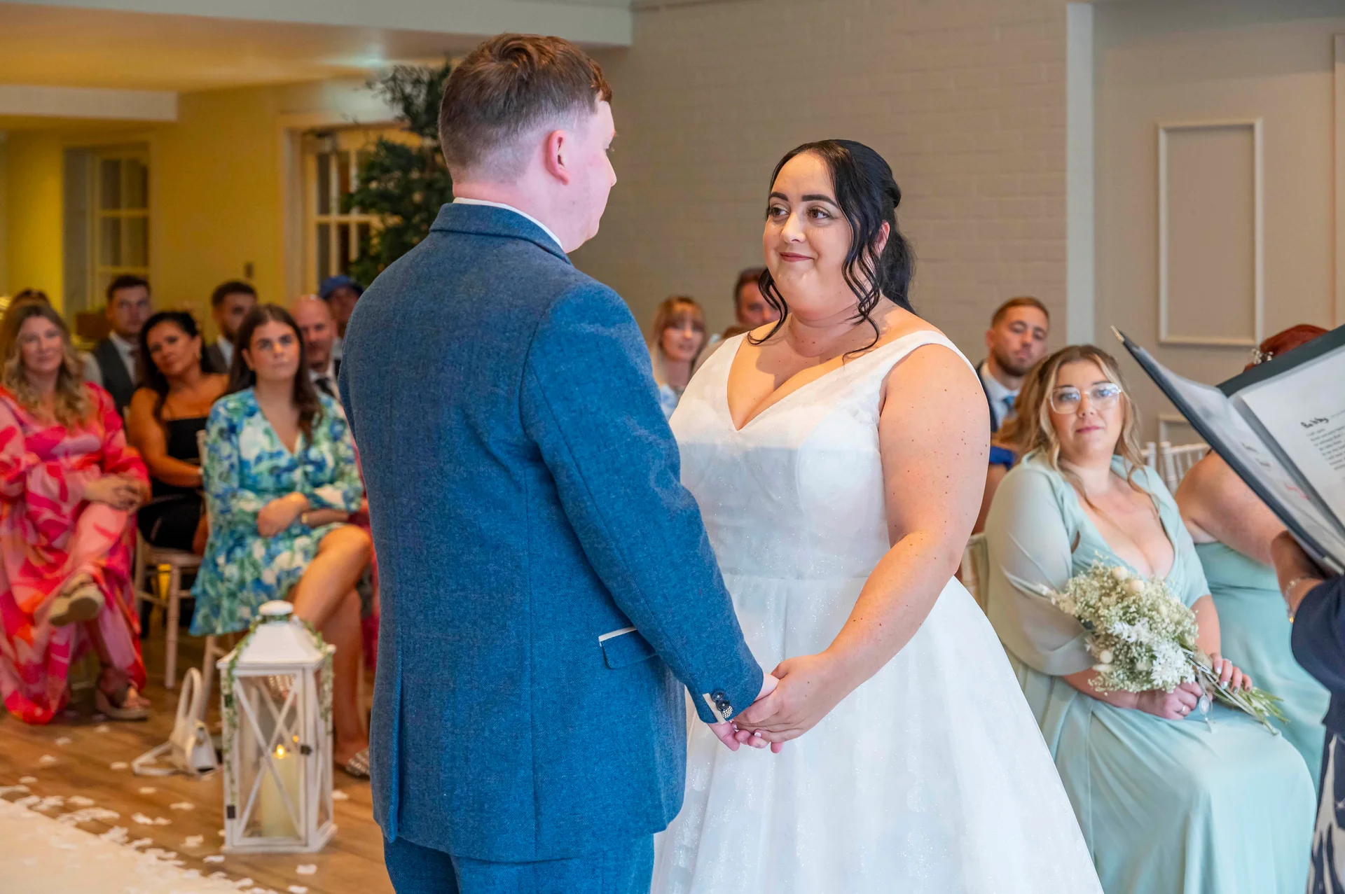 A groom in a blue suit and a bride in a white a-line dress hold hands while exchanging vows during an indoor wedding ceremony.