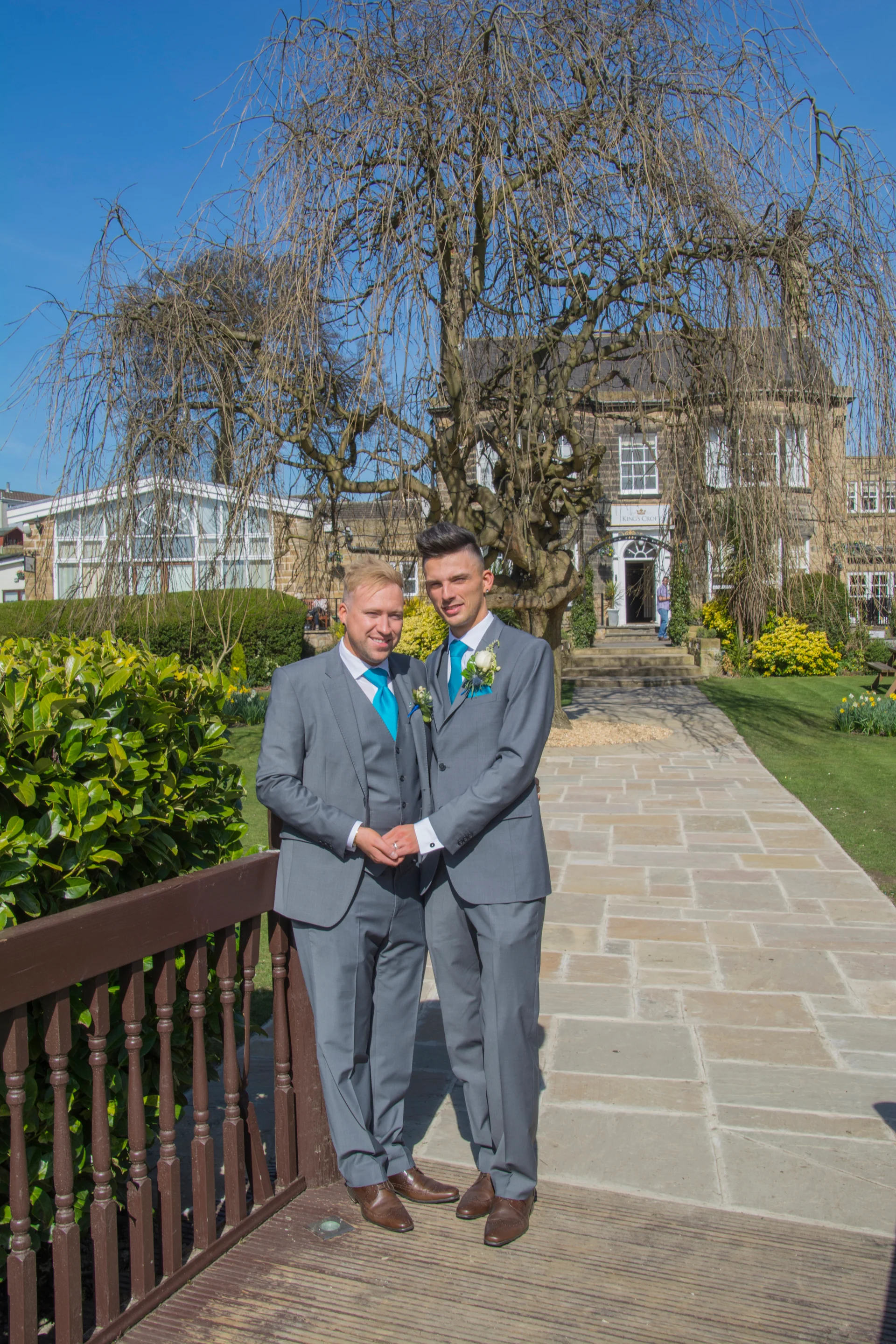 Two grooms in matching grey suits and blue ties stand holding hands on a wooden bridge during their hotel wedding.