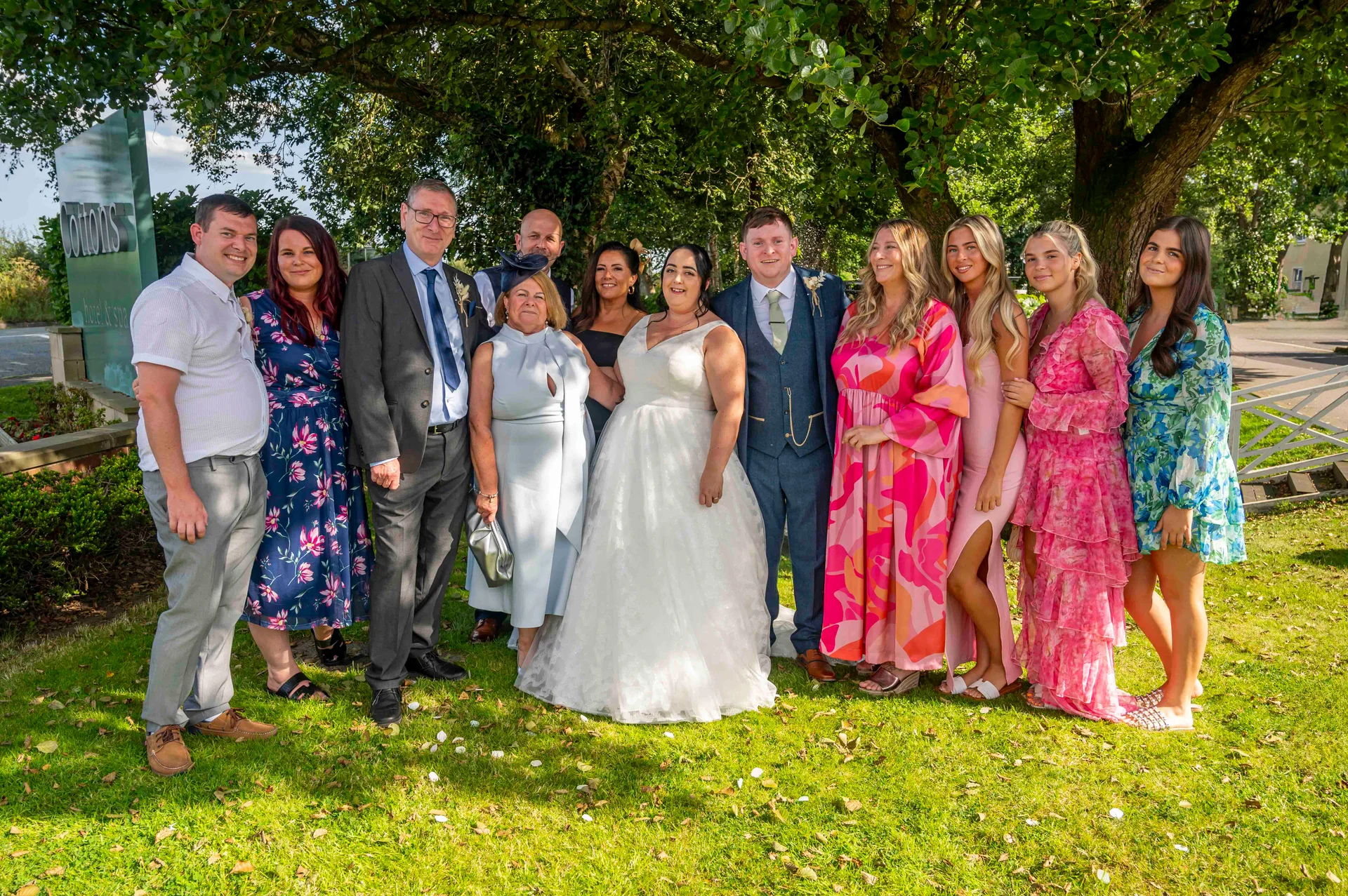 A newlywed couple poses with family and friends on a lush green lawn beneath a large leafy tree outside a hotel venue.