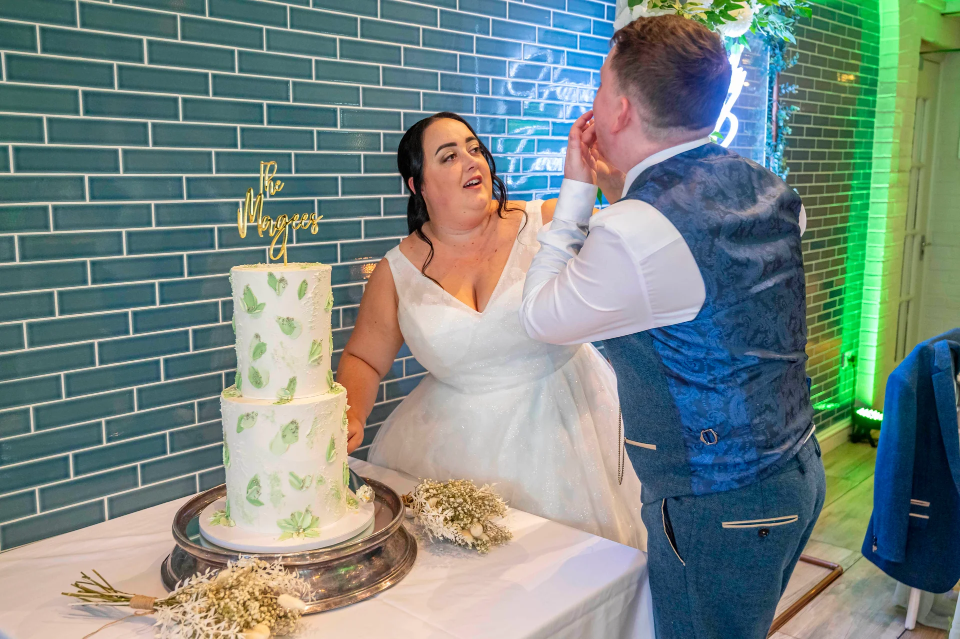 A bride and groom share a playful moment feeding each other wedding cake in front of a blue tiled wall.