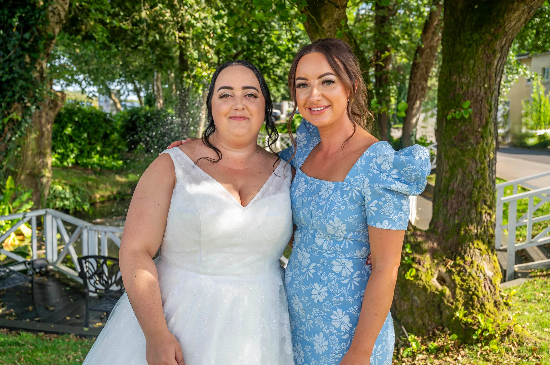A bride in a white A-line wedding dress poses outdoors alongside her partner who wears a blue patterned dress with puff sleeves.