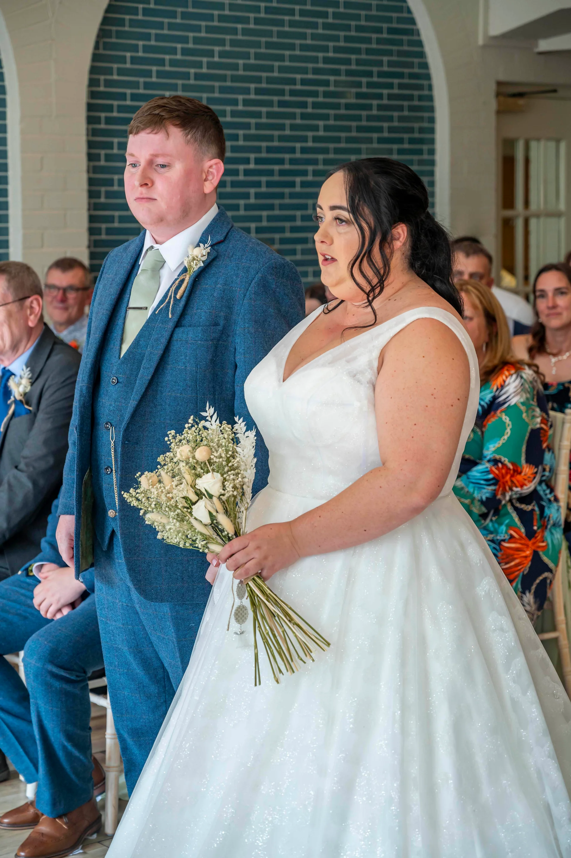 A newlywed couple stands together during their ceremony, with the bride holding a delicate bouquet and wearing a sparkling A-line gown while the groom wears a smart blue tweed suit.
