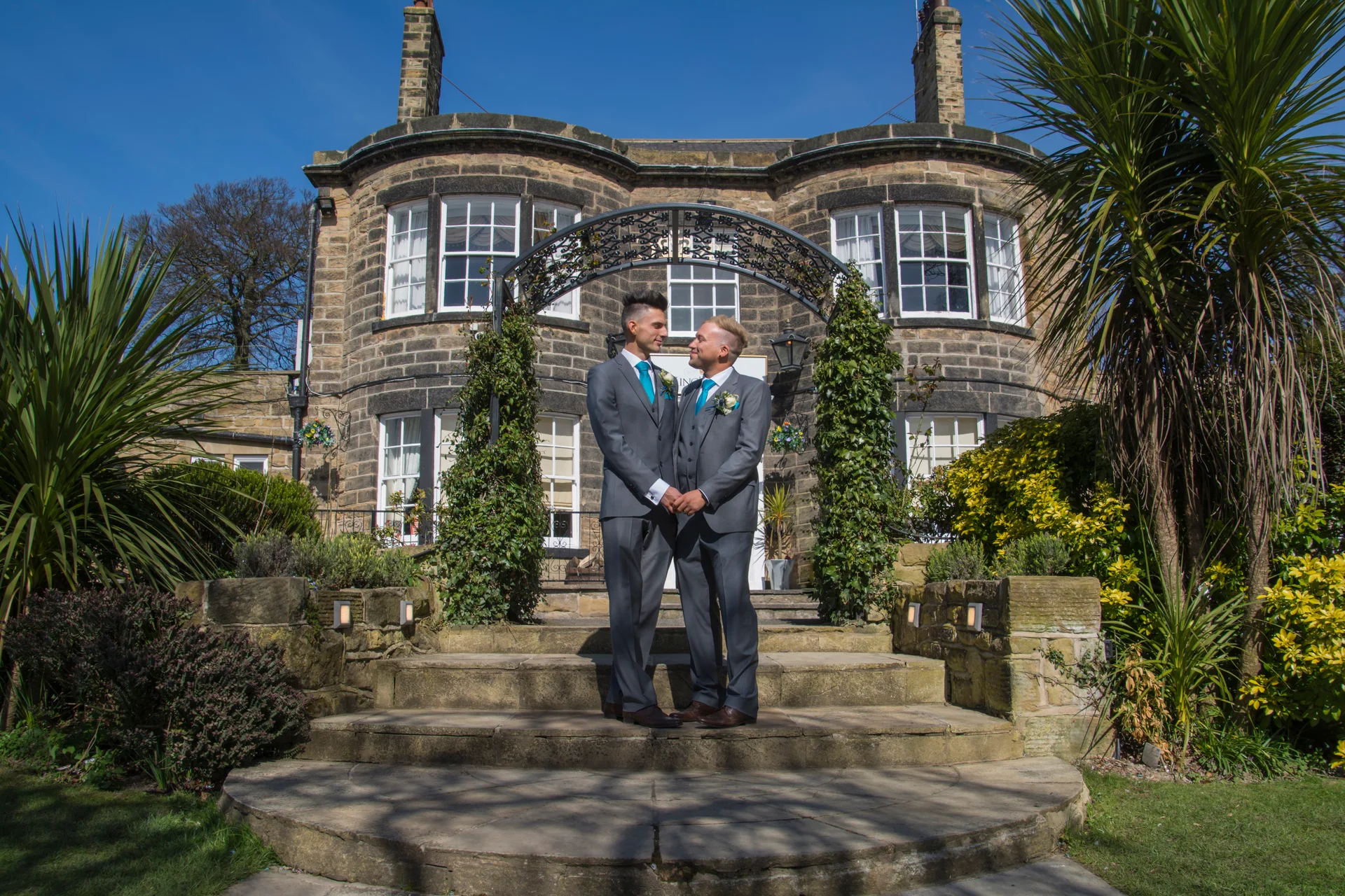 Two grooms in matching grey suits stand hand in hand beneath a floral archway on stone steps in front of a grand manor house under a clear blue sky.