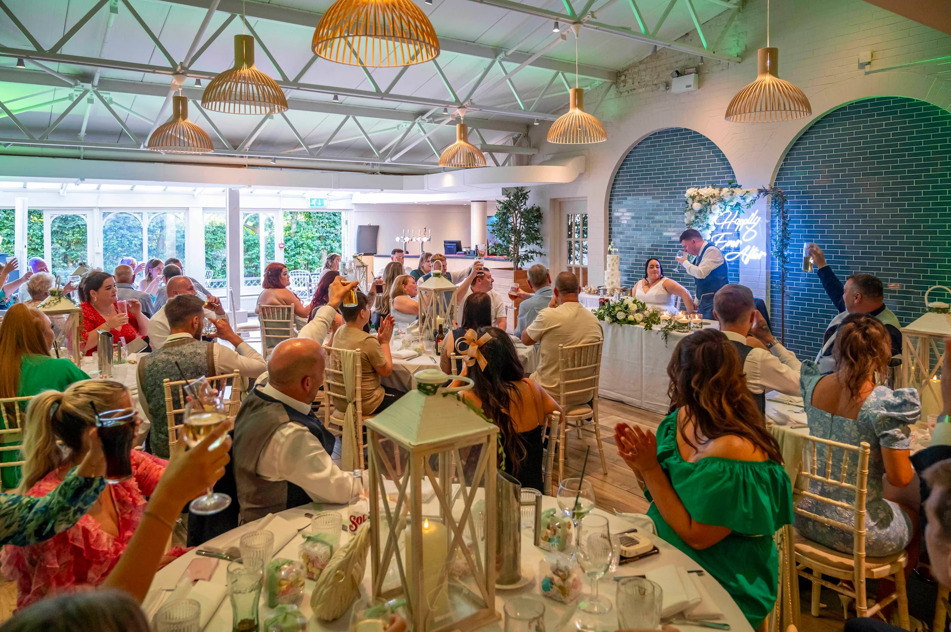Wedding guests raise their glasses in applause during speeches at a bright industrial venue featuring exposed rafters and modern pendant lighting.