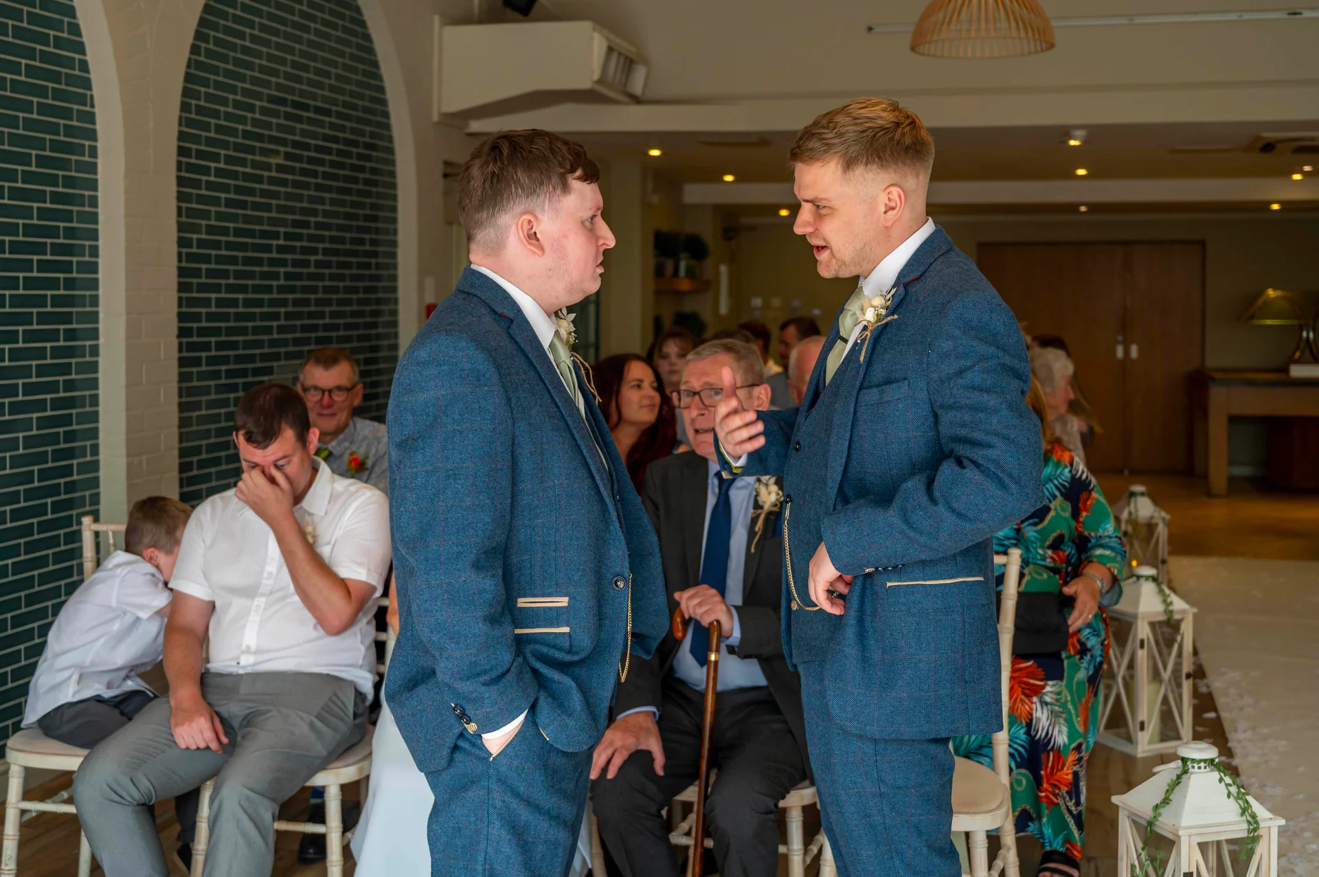 Two grooms in matching blue tweed suits converse during their wedding ceremony as seated guests look on and one guest reacts emotionally.