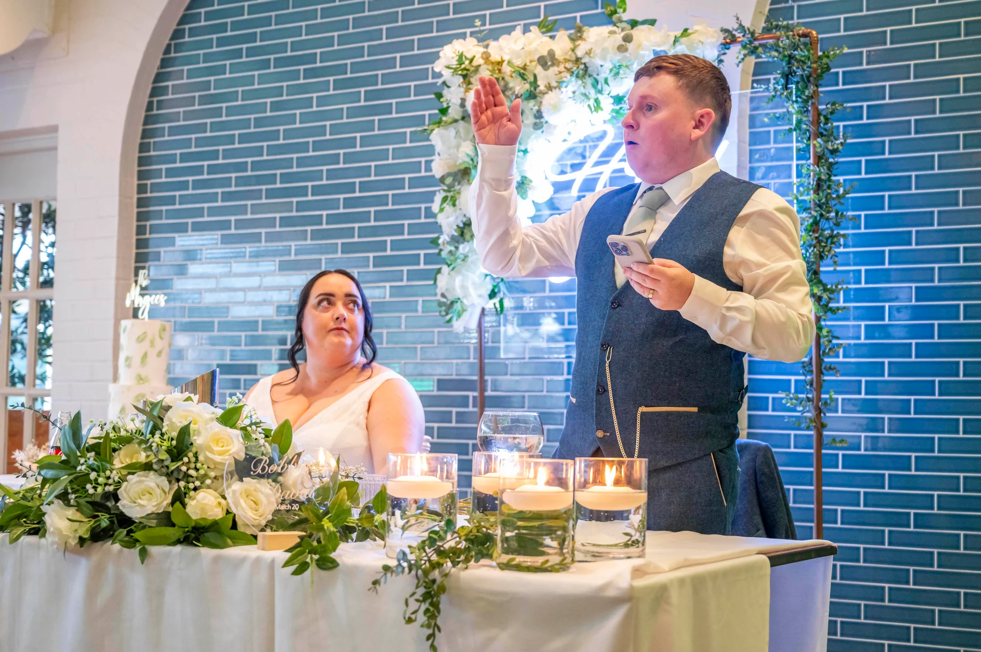 A groom delivers a speech holding a phone while standing beside his seated bride at the top table, set against a teal tiled wall.