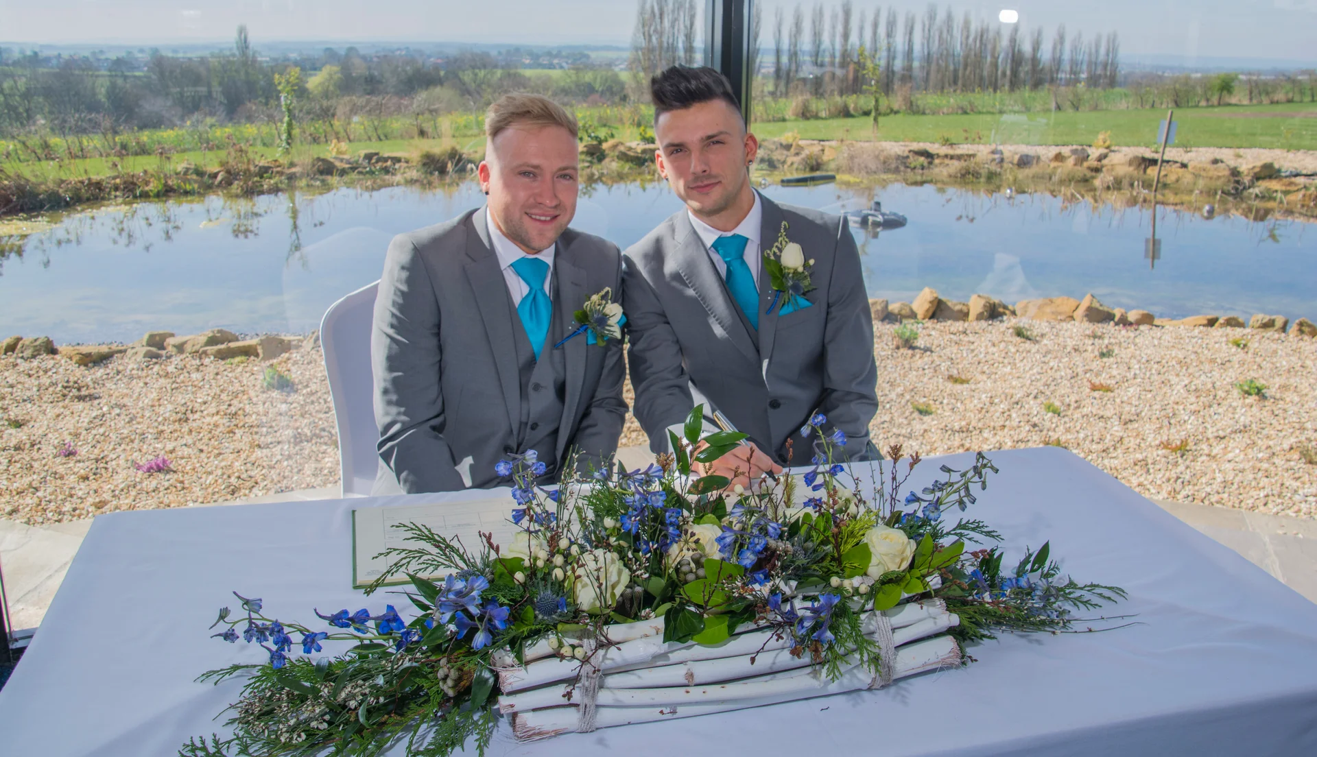 Two grooms smile for the camera while signing the register at a table adorned with floral arrangements, with a scenic lake visible through the large window behind them.