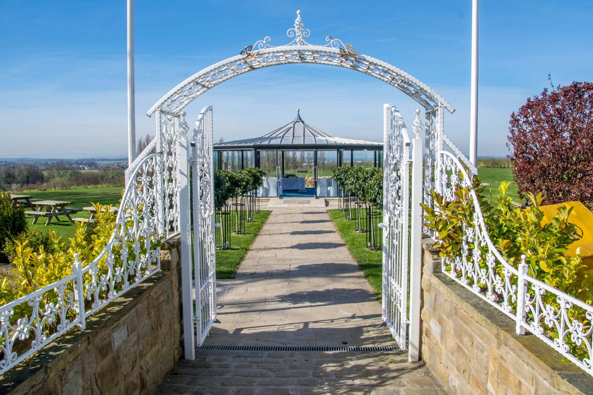 An ornate white iron archway frames a paved stone path leading towards a glass gazebo set against a bright blue sky and rolling green hills.