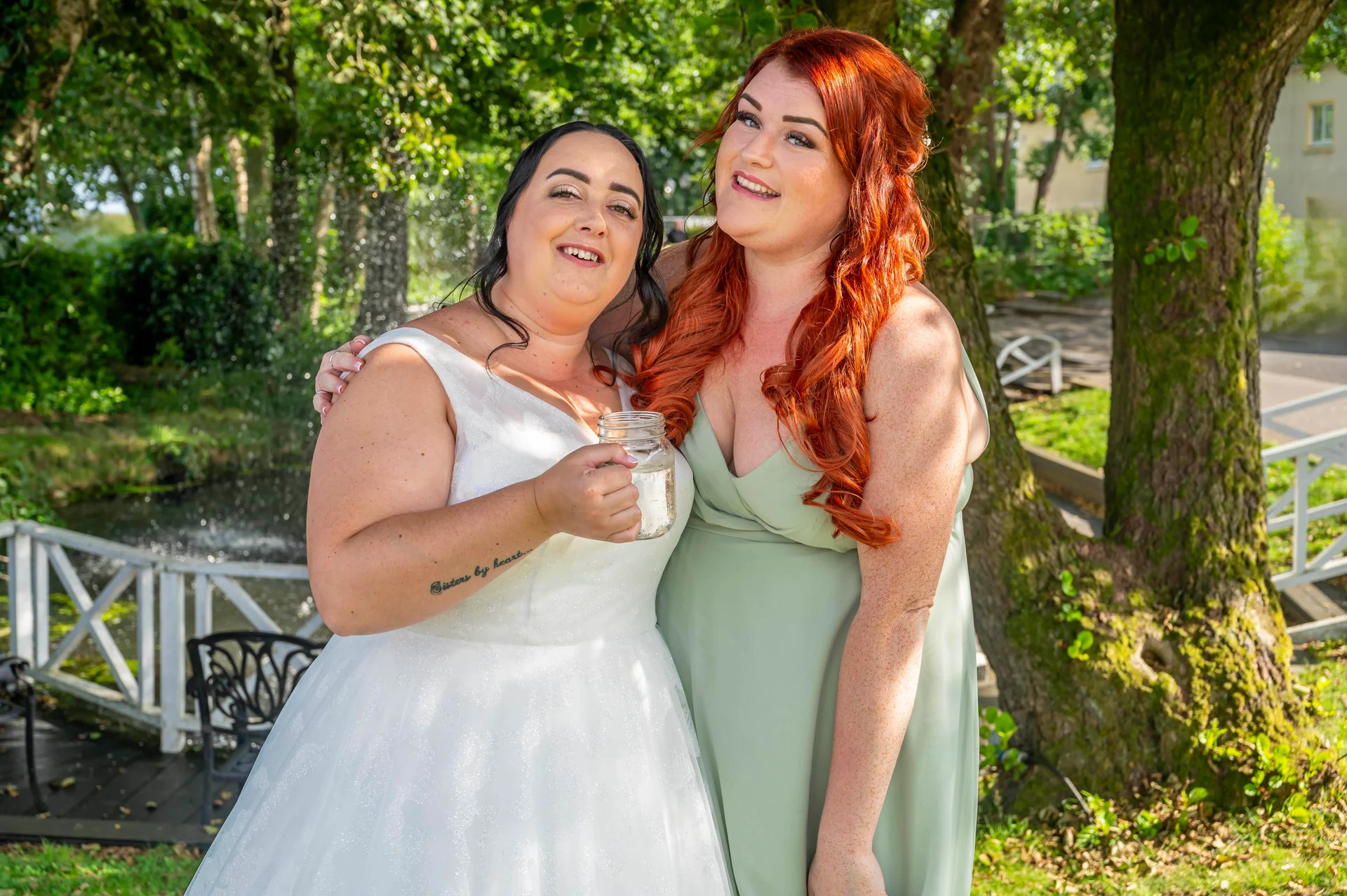 Two smiling brides embrace outdoors in a sunlit garden beside a reflective pond, with one wearing a sparkling white a-line gown and the other in a sage green dress.