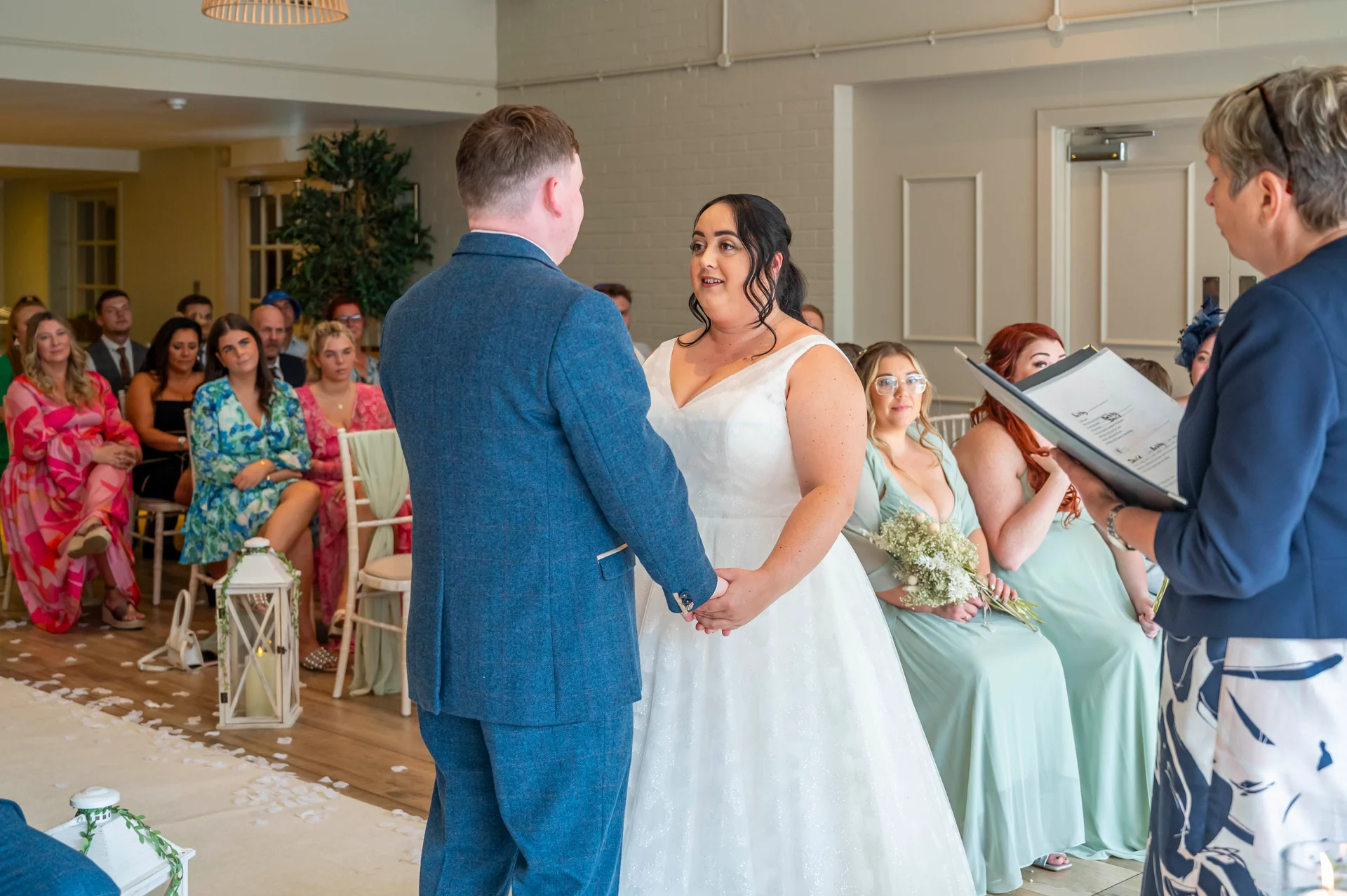 A bride in a white a-line gown and a groom in a blue tweed suit hold hands during their civil wedding ceremony while guests watch.