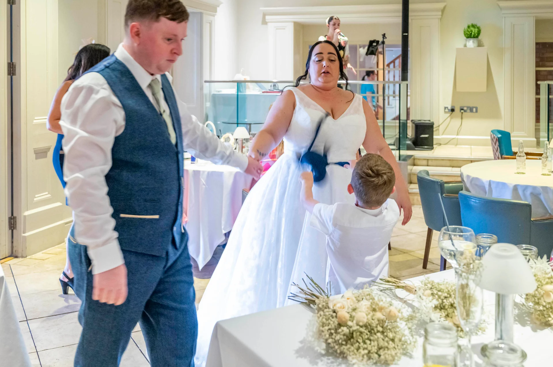 A groom in a blue tweed waistcoat and a bride in a white A-line dress walk through a reception venue while a young boy reaches toward the bride's dress near a table adorned with floral centrepieces.