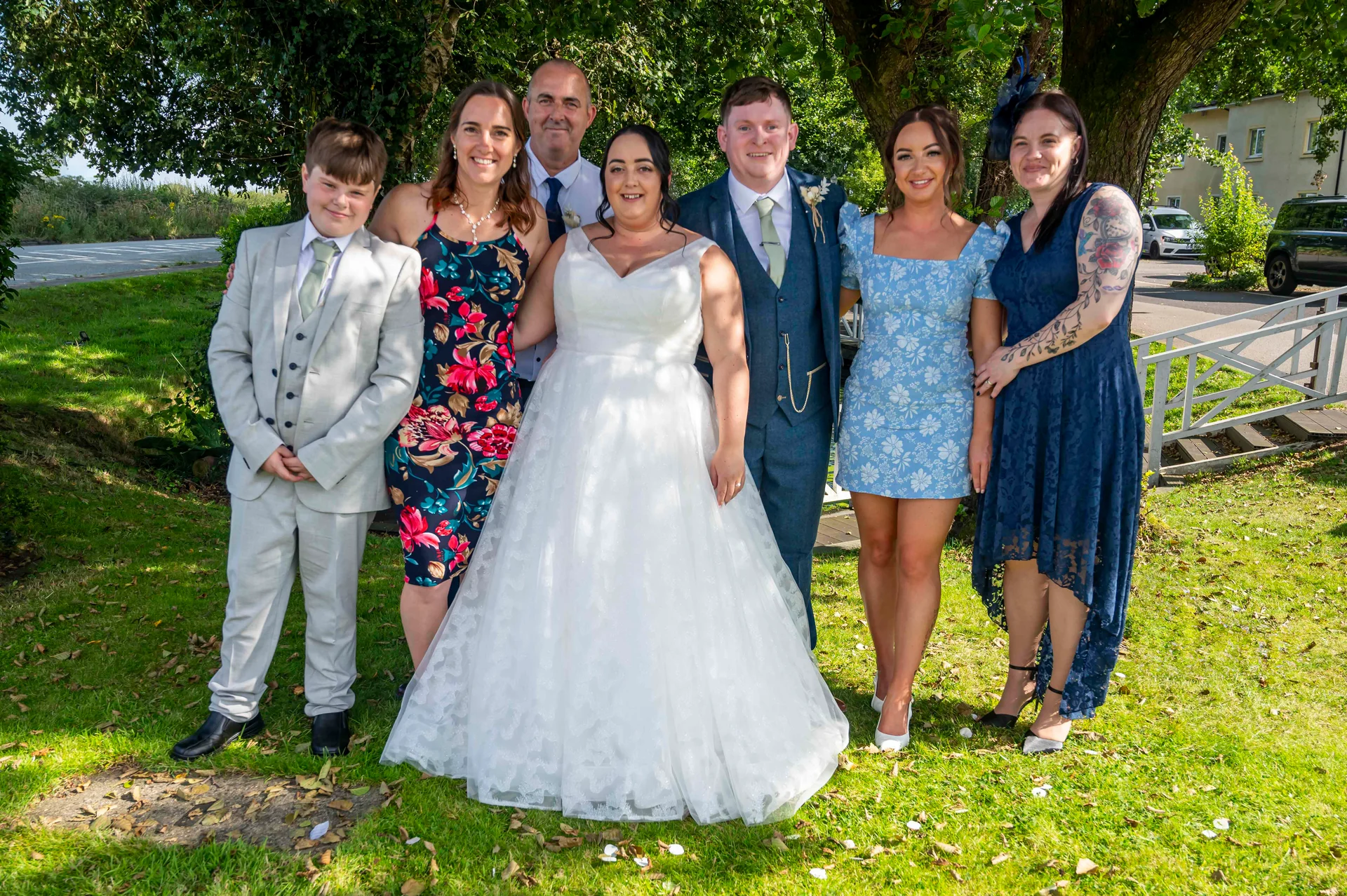 A happy wedding party poses outdoors on a sunlit lawn near trees, featuring the bride in a white gown and the groom in a blue suit alongside family and friends.