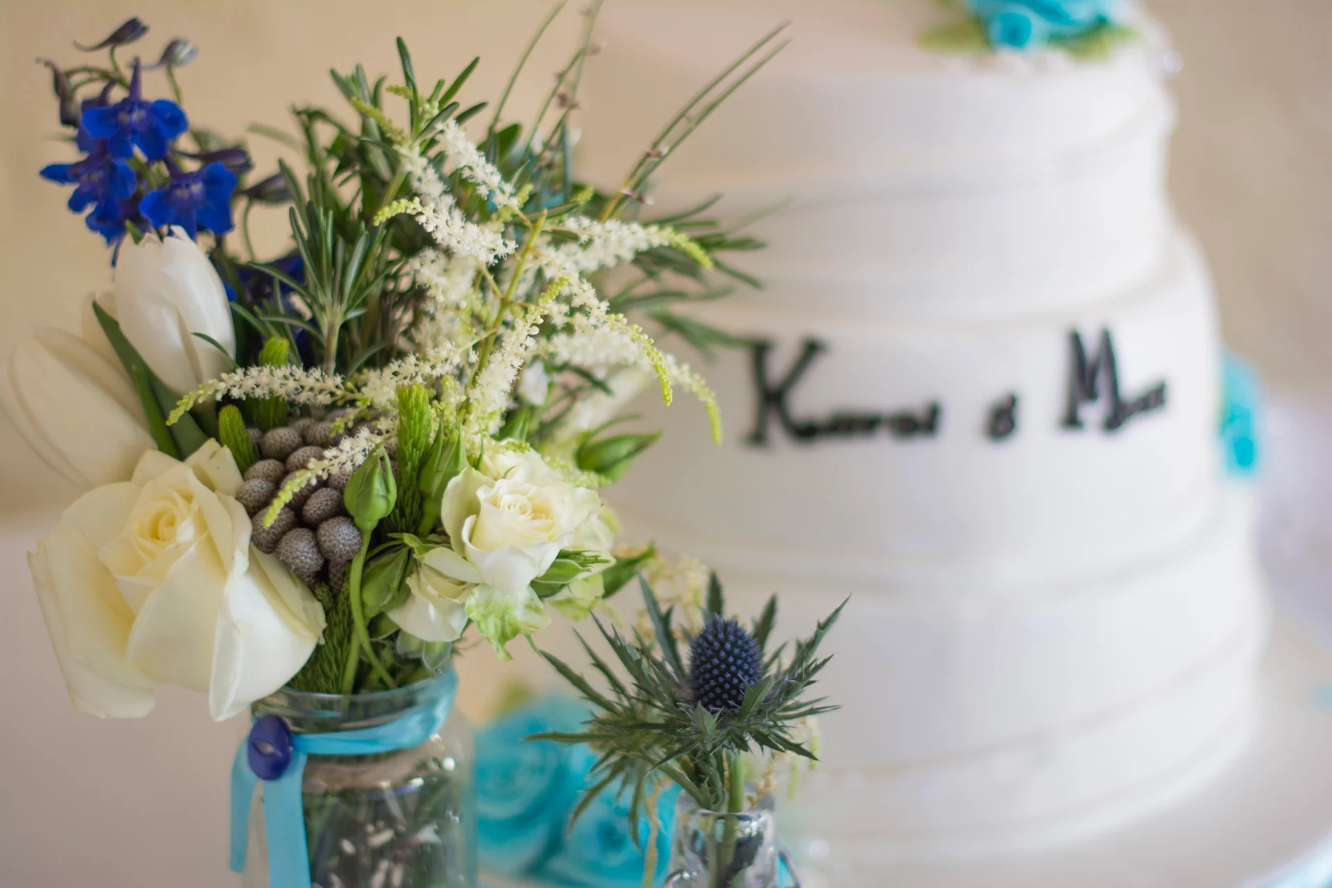A rustic wedding cake featuring white tiers and blue floral accents sits behind a glass jar filled with white roses and blue delphiniums.