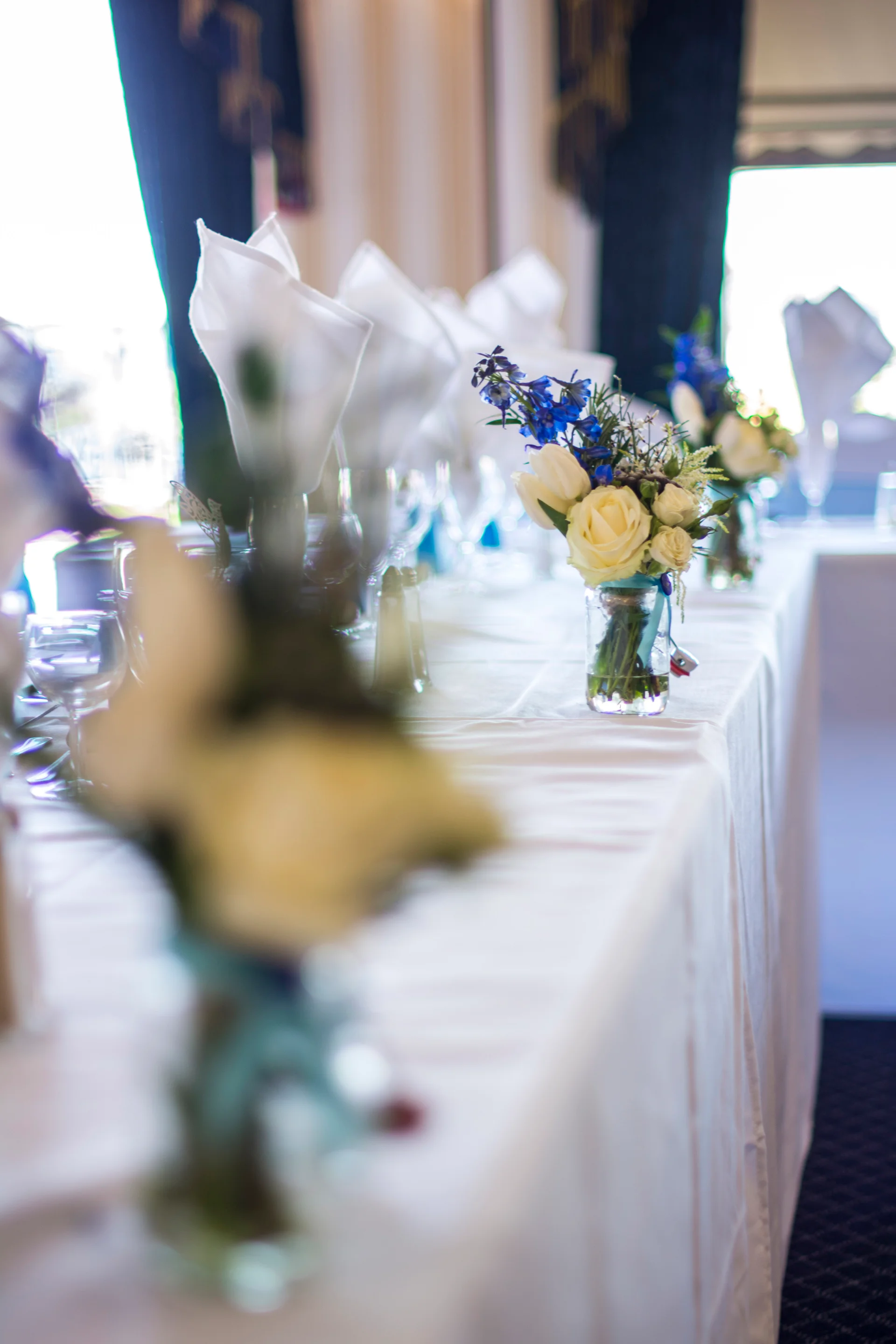 An elegant wedding top table is adorned with crisp white linen, neatly folded napkins, and delicate bud vases filled with white roses and blue delphiniums, bathed in soft window light.