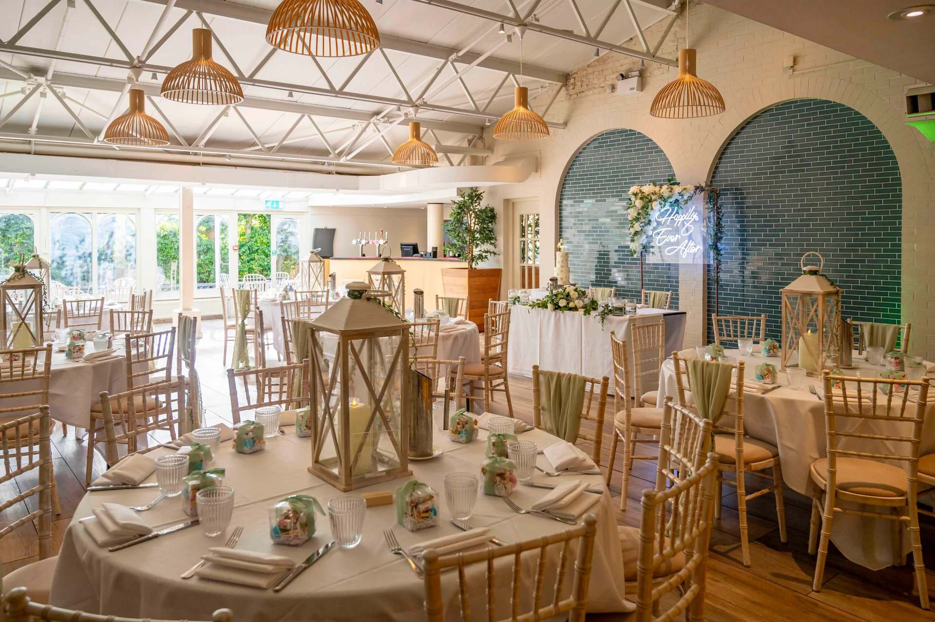 An elegantly decorated wedding reception venue featuring round tables with white linens, sage green napkins, wooden lanterns, and wooden chairs beneath a vaulted industrial ceiling.