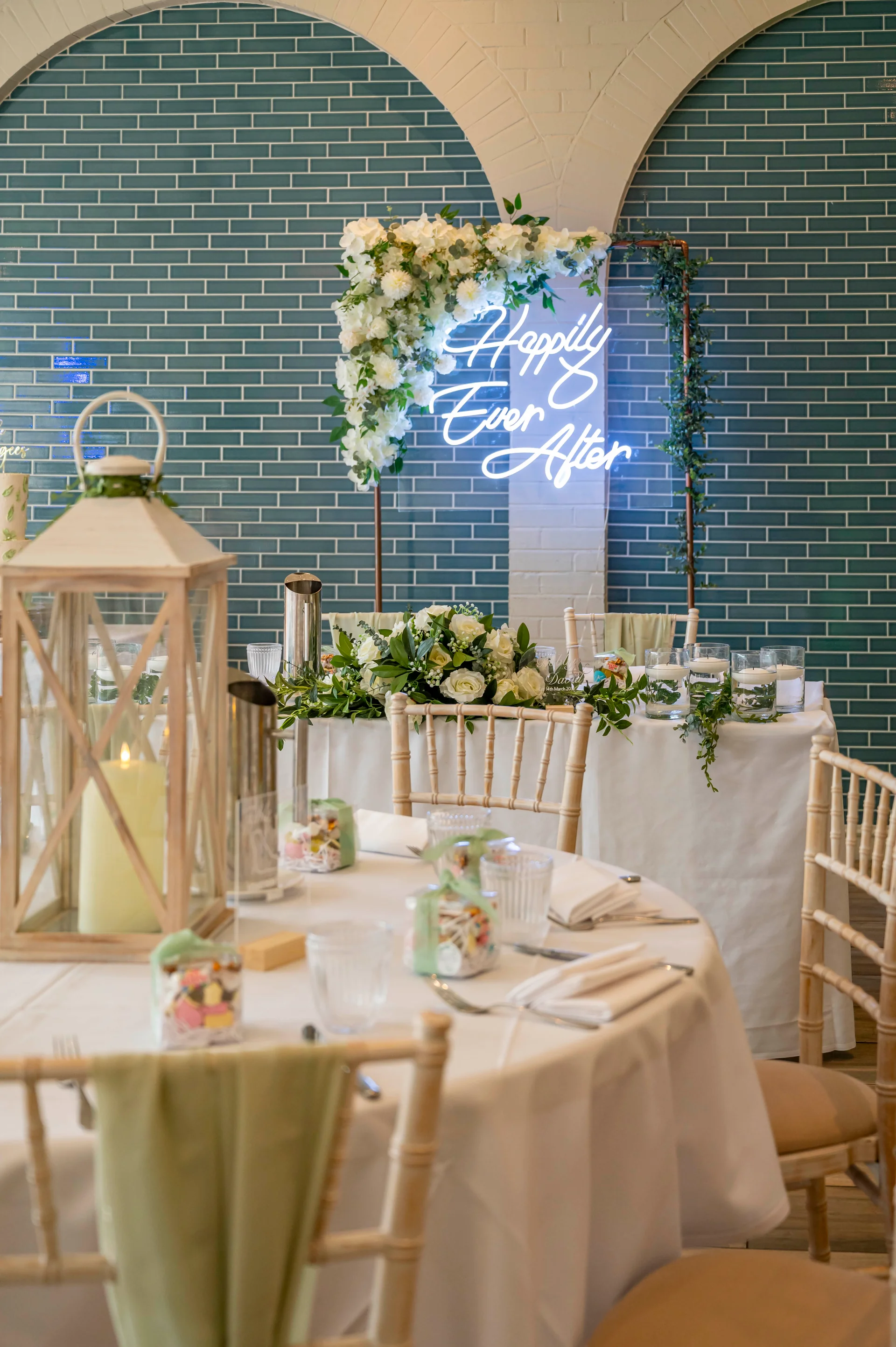 A beautifully styled wedding reception venue featuring round tables with white linens, chiavari chairs, and a top table decorated with a white floral arrangement and an illuminated neon sign against a teal tiled wall.