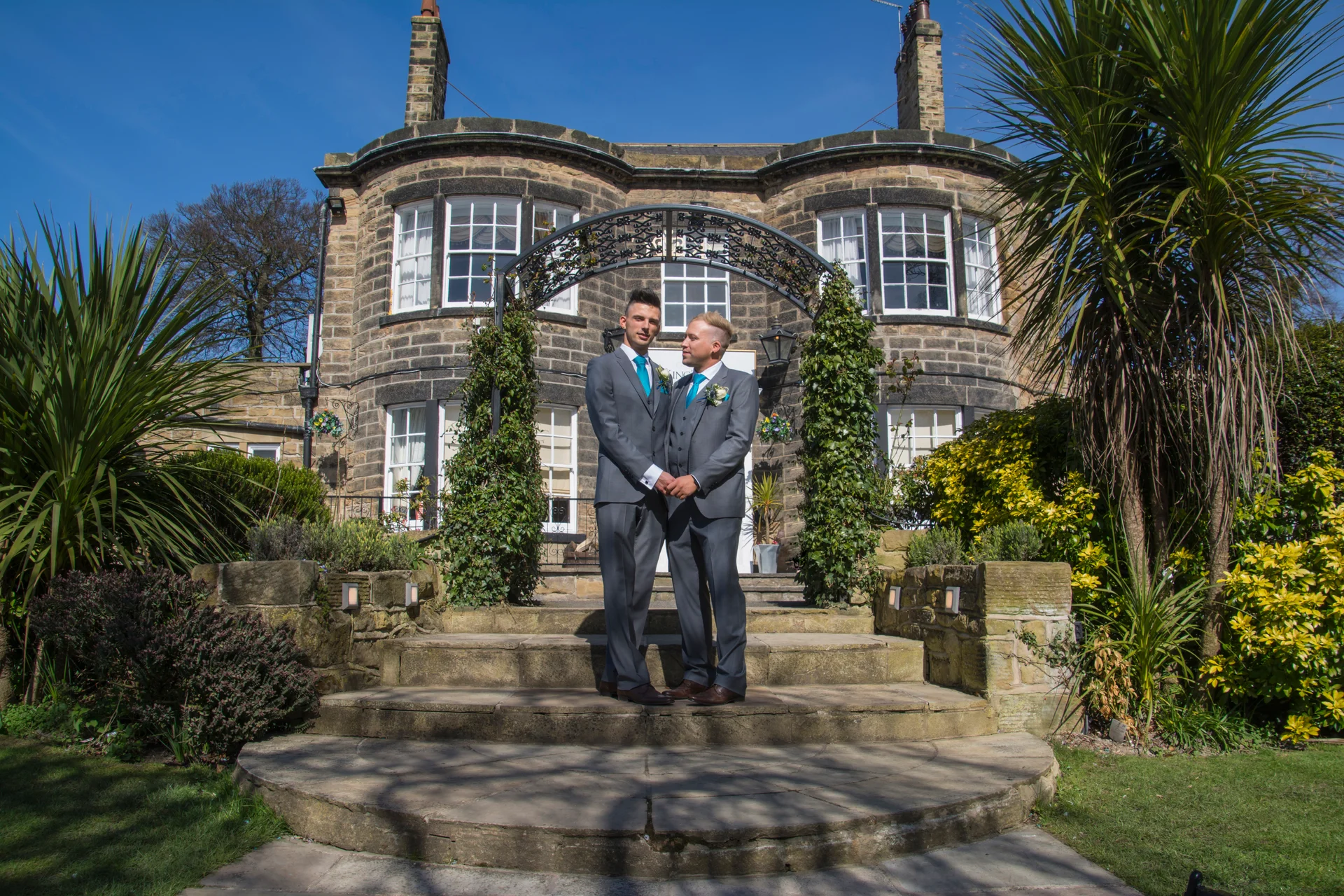 Two grooms in matching grey suits stand hand in hand on stone steps outside a grand manor house under a clear blue sky.