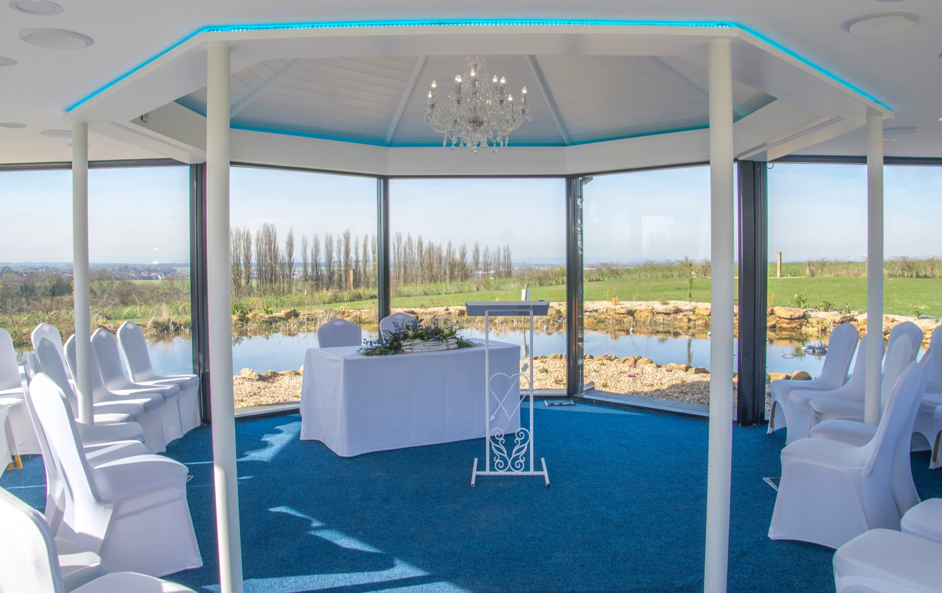An empty modern ceremony room featuring floor-to-ceiling panoramic windows overlooking a serene lake and countryside, set up with white-covered chairs and a signing table under a crystal chandelier.