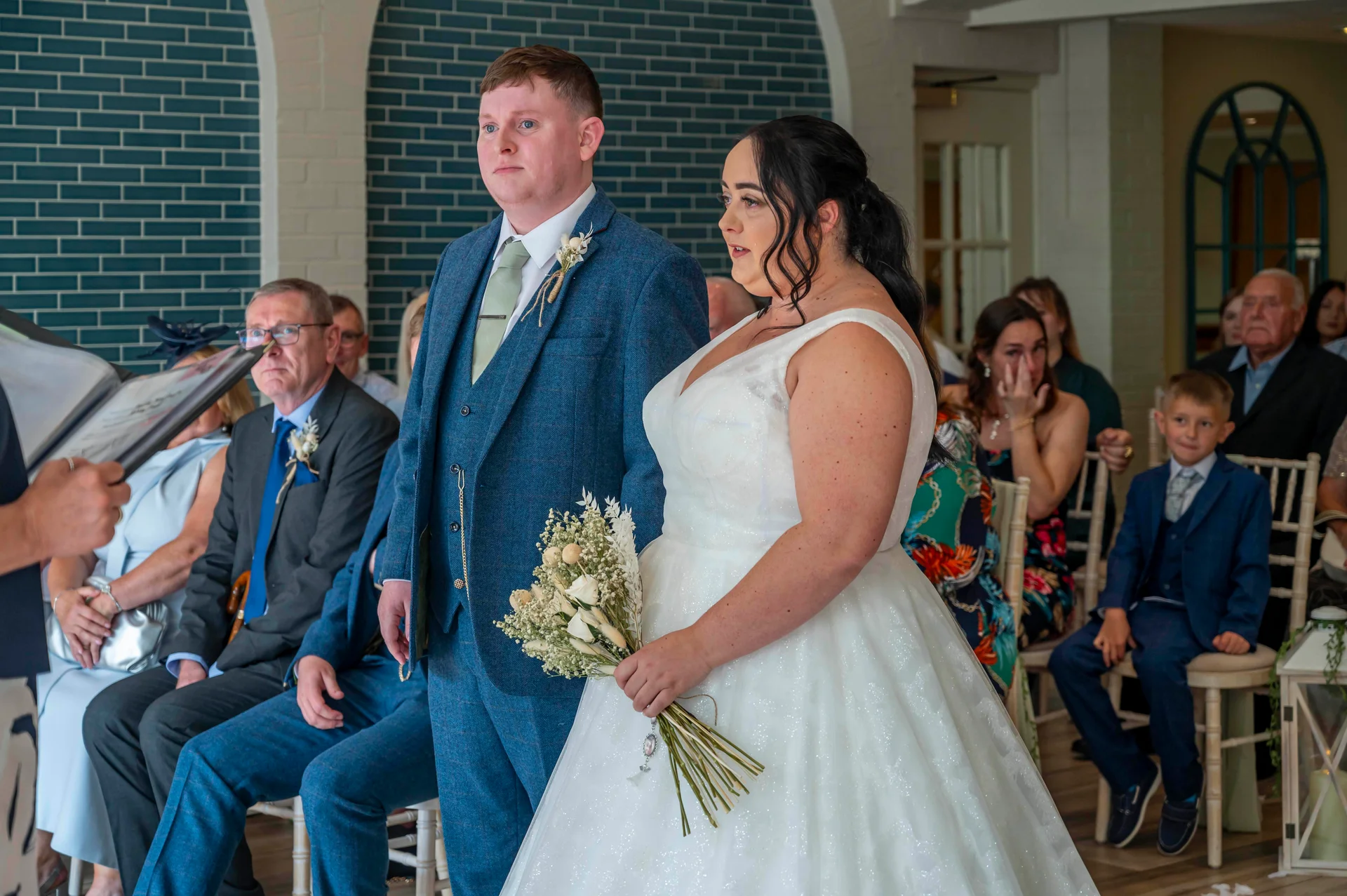 The bride holding a dried flower bouquet stands next to the groom in a blue tweed suit during their indoor wedding ceremony while guests watch emotionally.