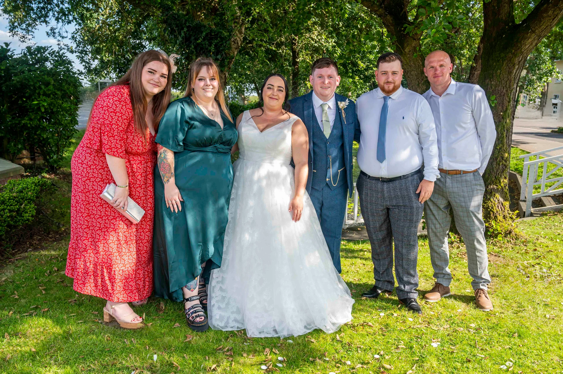 A bridal party poses outdoors on a grassy lawn beneath shady trees on a bright sunny wedding day.
